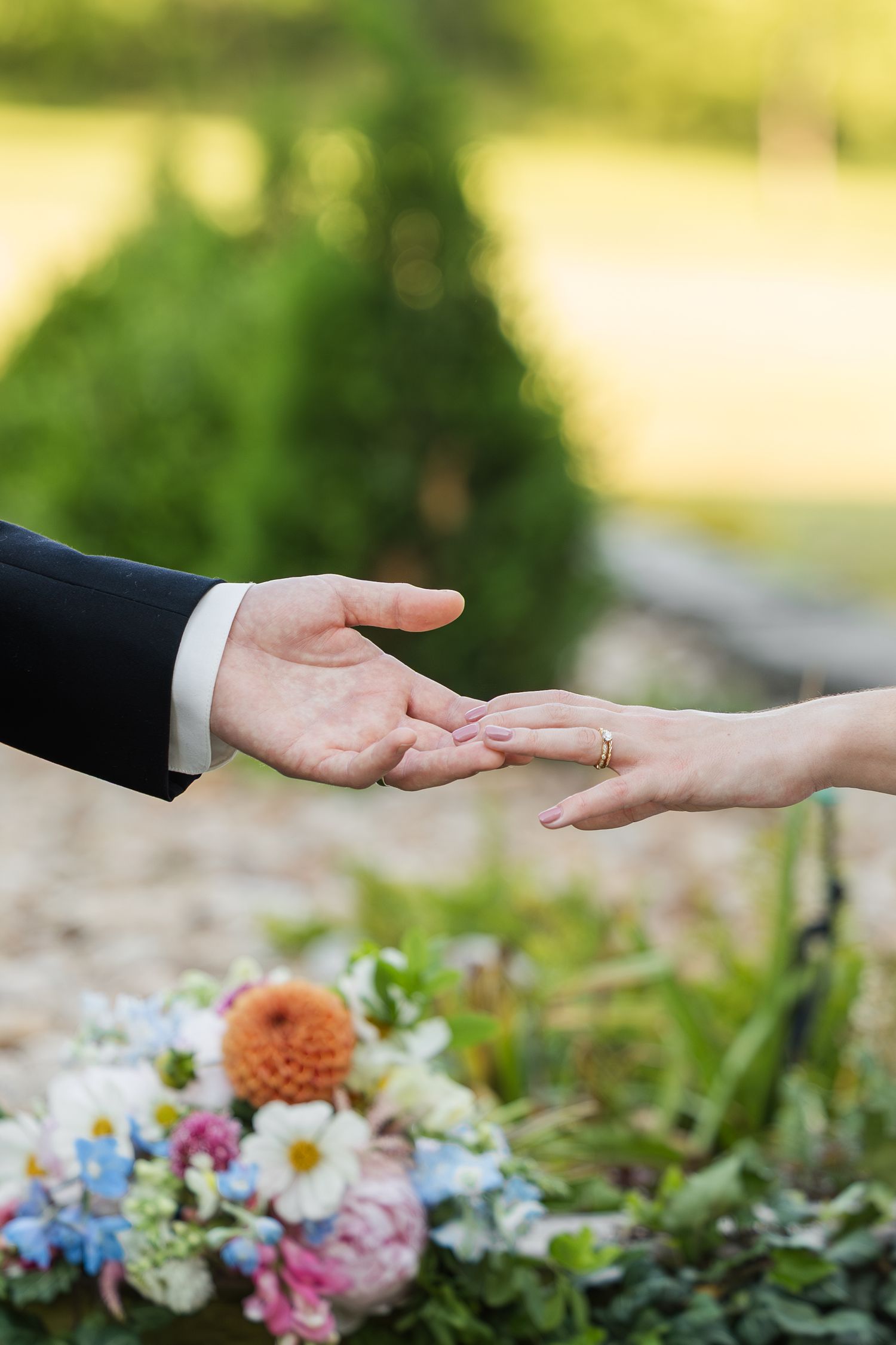 Bride and groom gently touching hands outdoors, with a colorful wedding bouquet of dahlias and daisies in the foreground.
