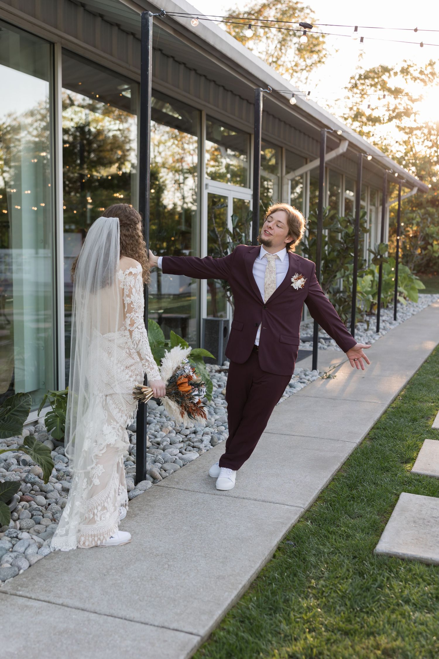Bride in lace gown and veil meeting partner in burgundy suit outside modern glass venue.