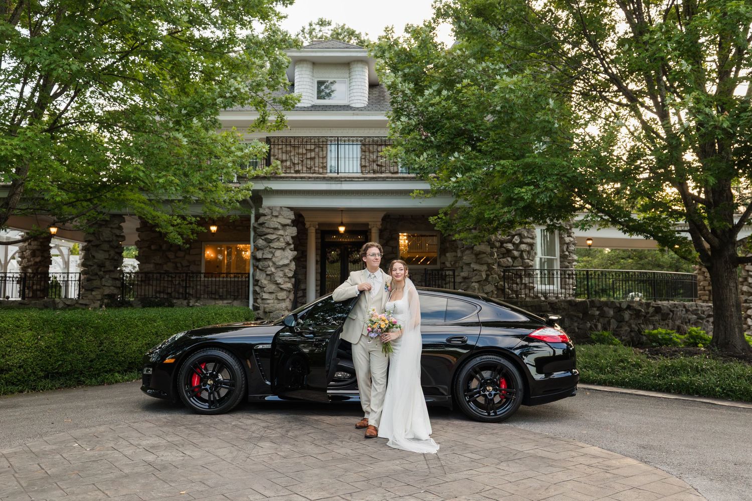 Bride and groom posing in front of a black Porsche at an elegant stone mansion wedding venue.