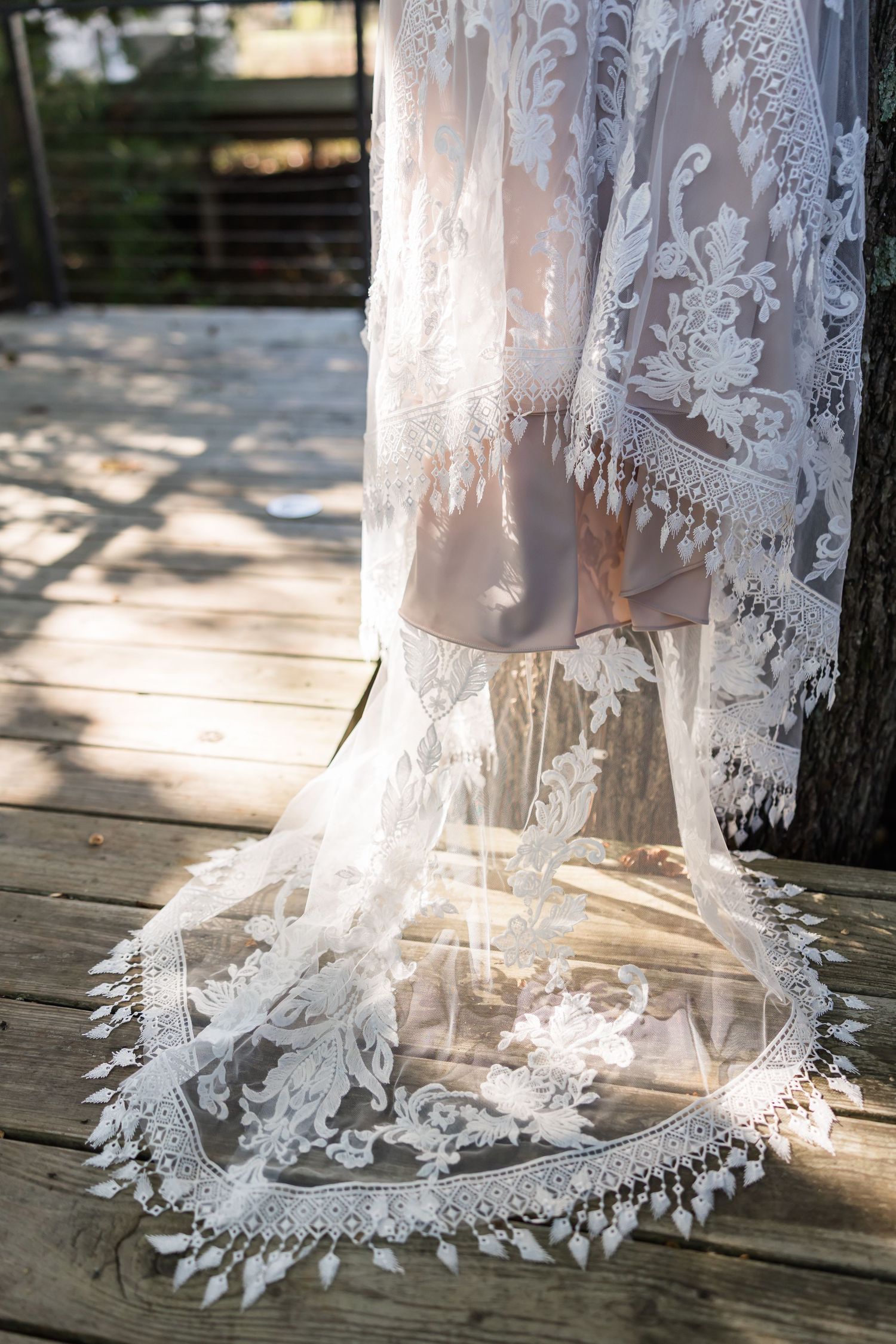 Elegant lace wedding veil with intricate floral patterns and fringe trim draped on a wooden deck in sunlight.
