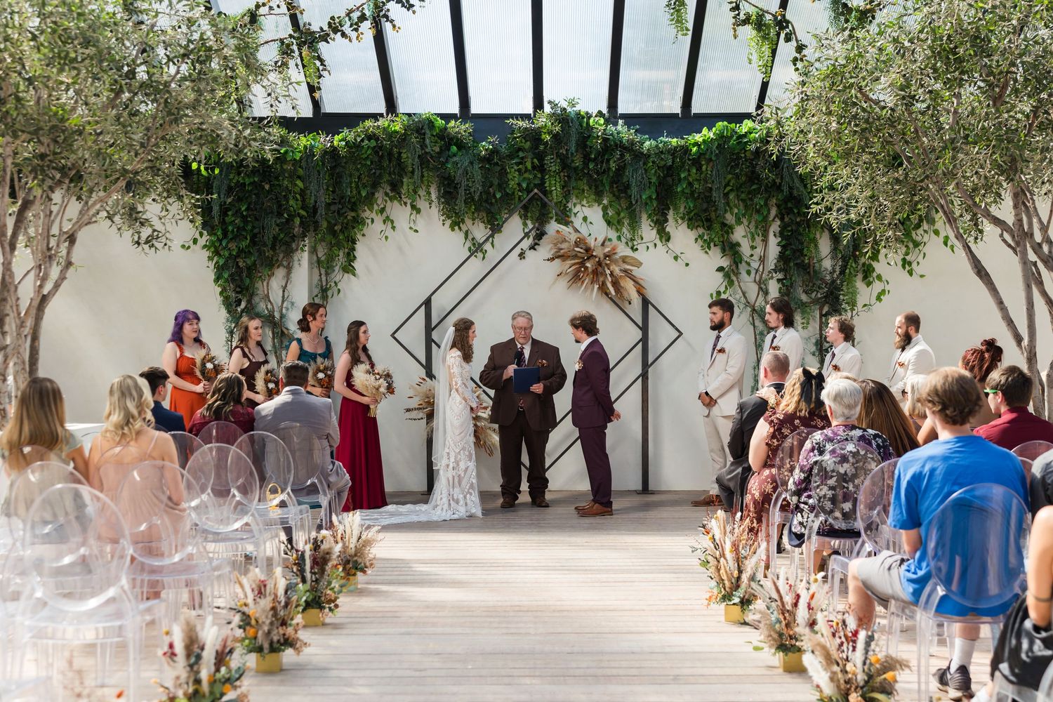 Elegant indoor wedding ceremony beneath lush greenery arch with guests seated in clear chairs.