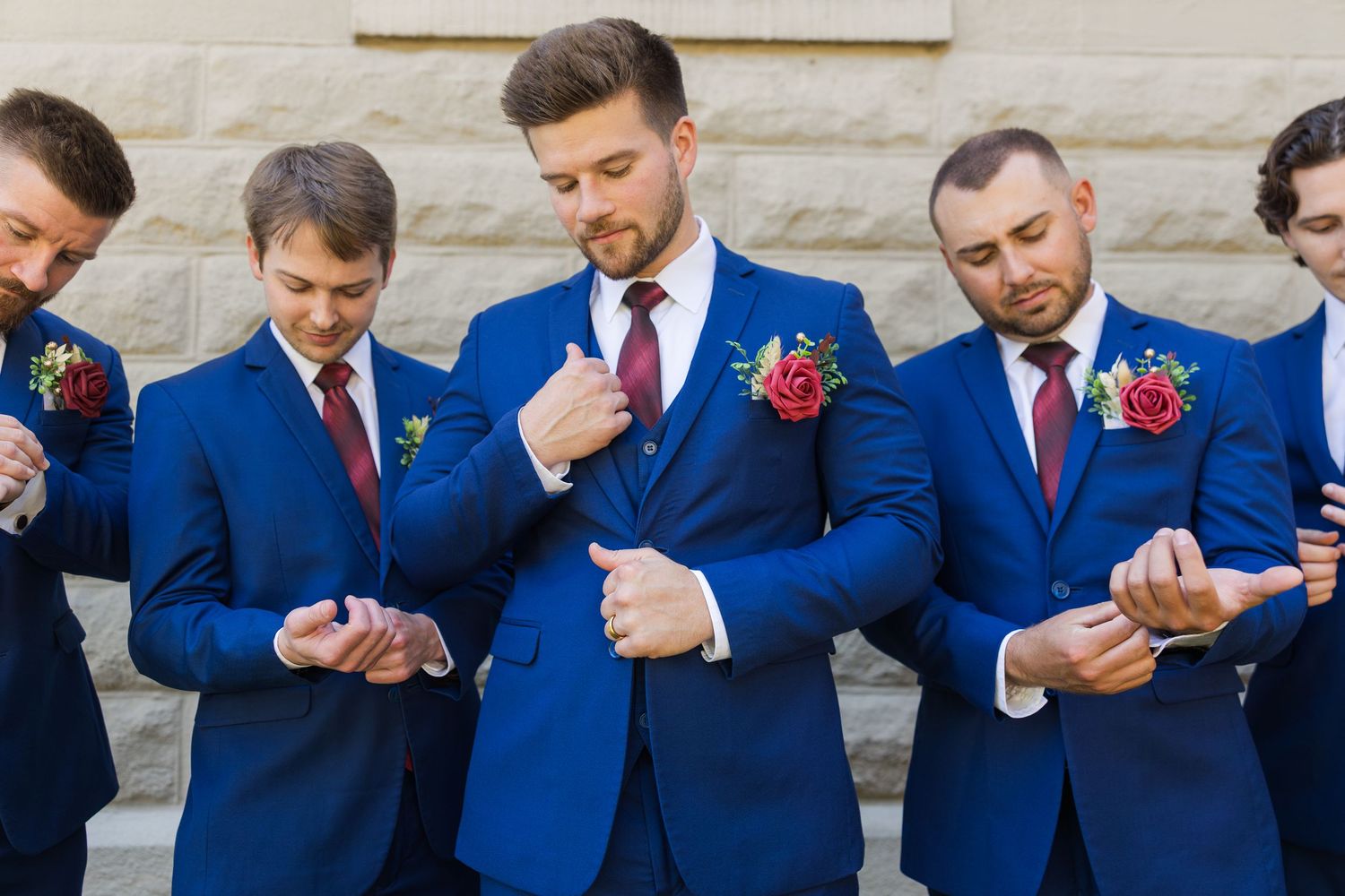 Five groomsmen in navy blue suits with burgundy boutonnieres adjust their cufflinks before a wedding ceremony.