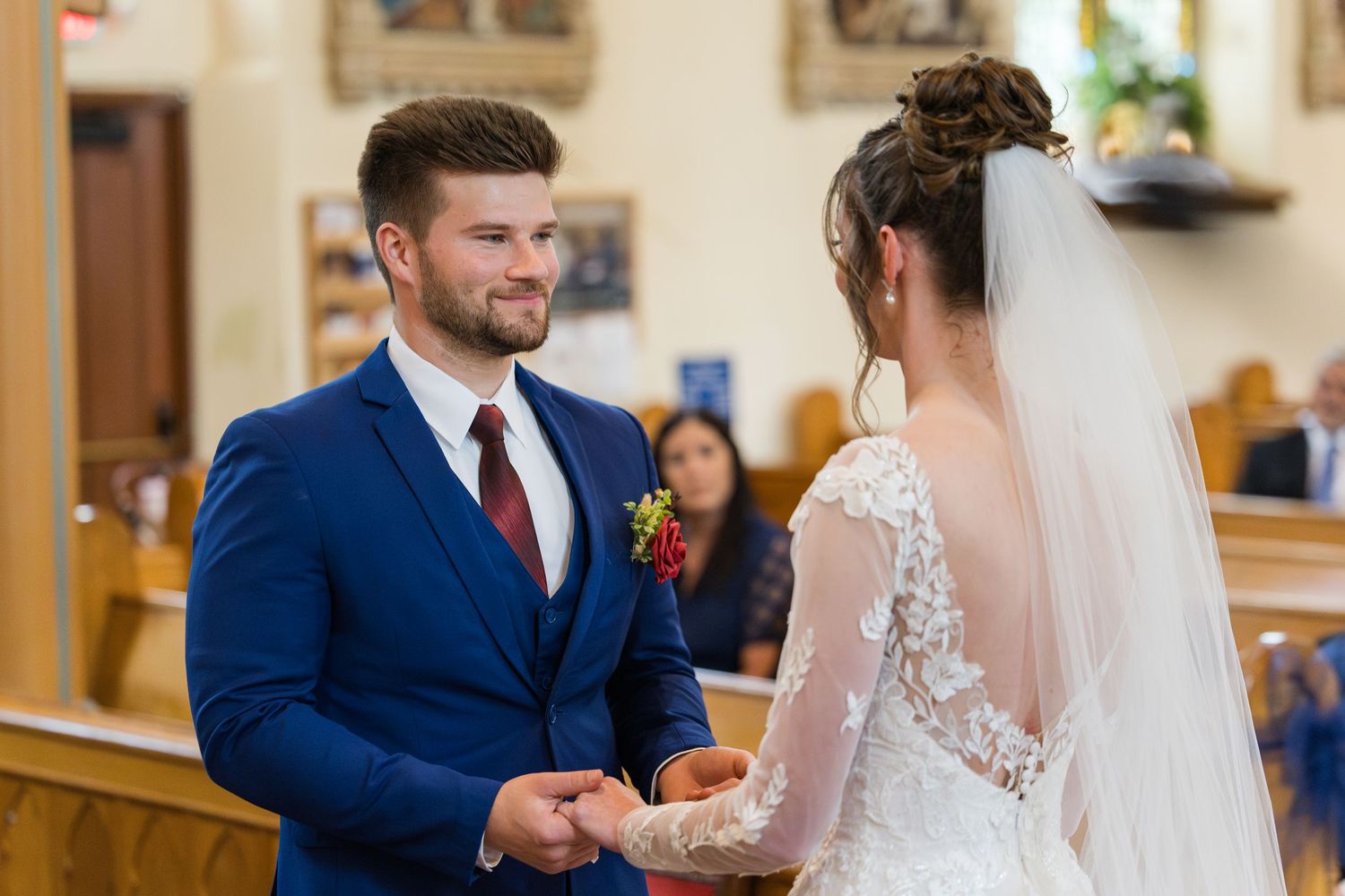 Groom in blue suit exchanges vows with bride in lace wedding dress during church ceremony.