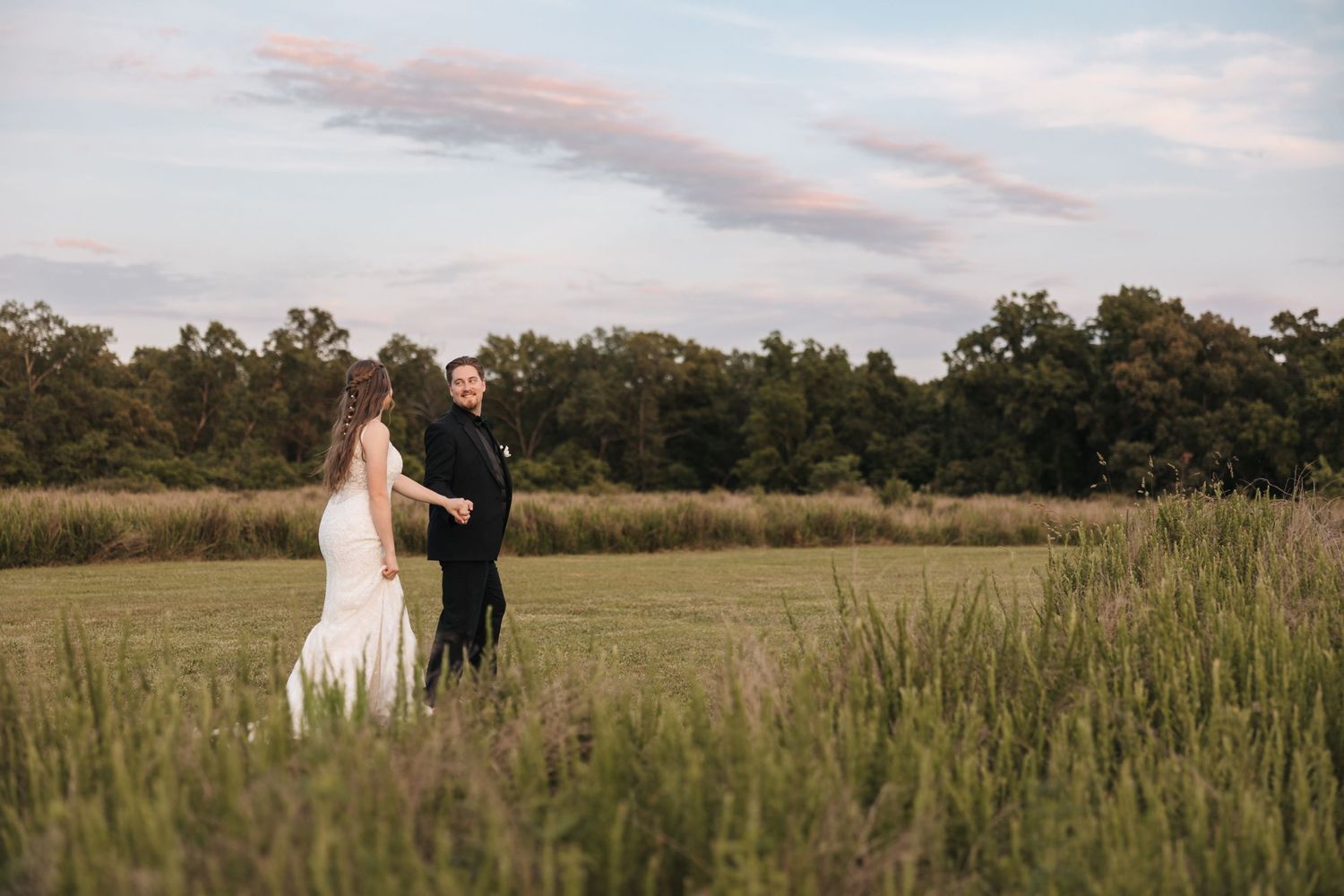 A couple in wedding attire stands together in a golden field at sunset.