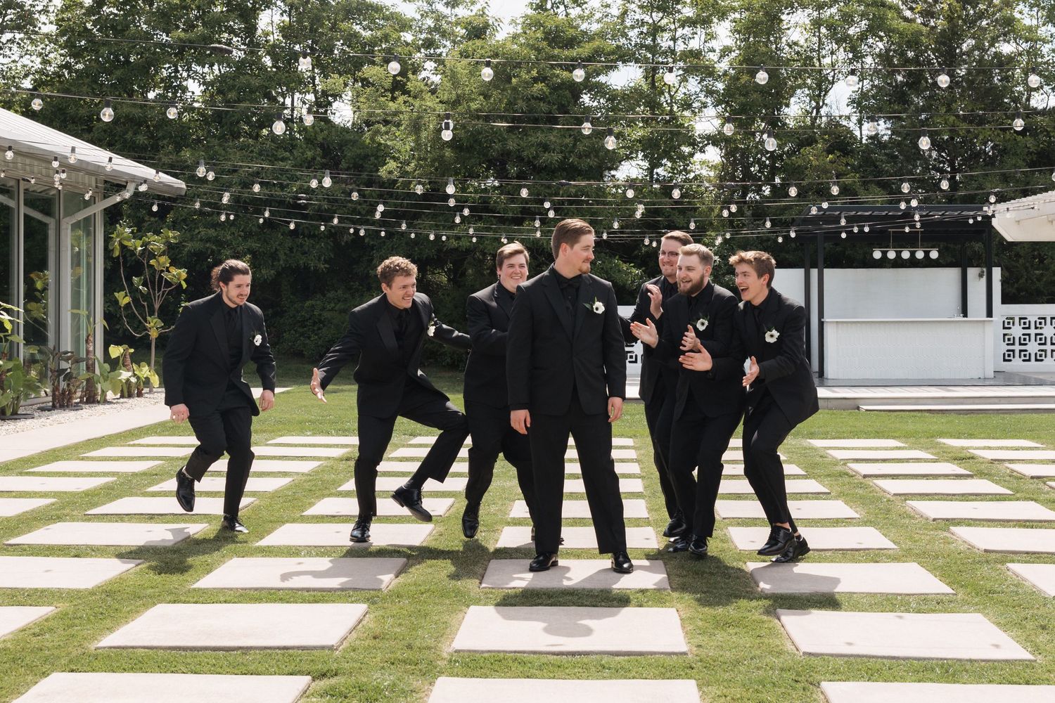 Groomsmen in black suits perform a fun jumping action shot on a geometric patio.
