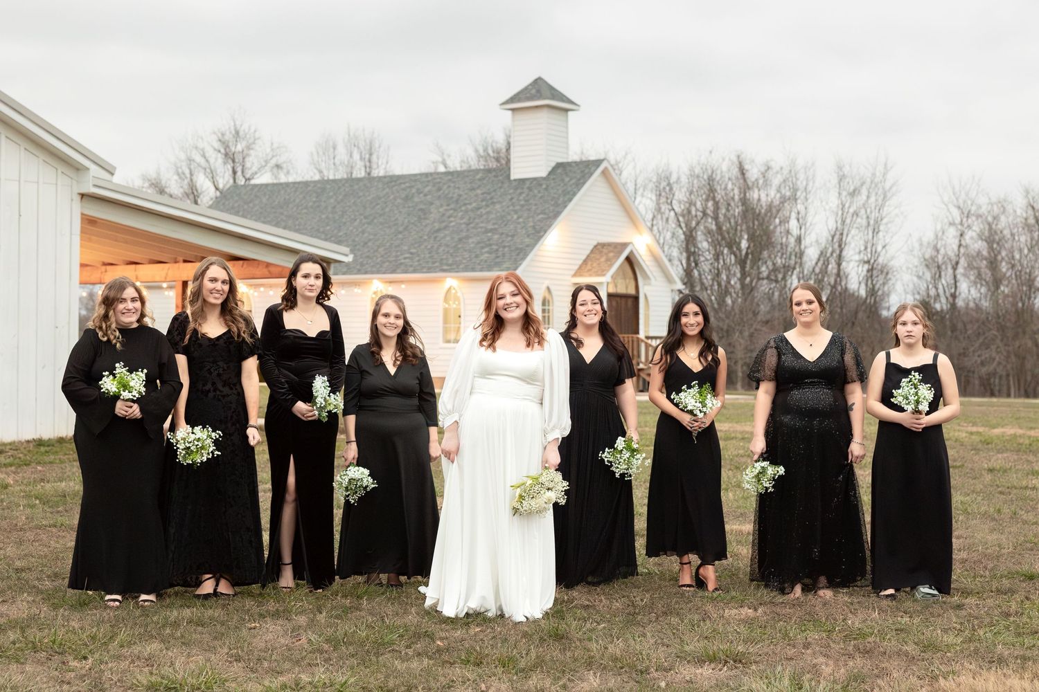 Wedding party in black dresses poses formally in front of a white chapel building.