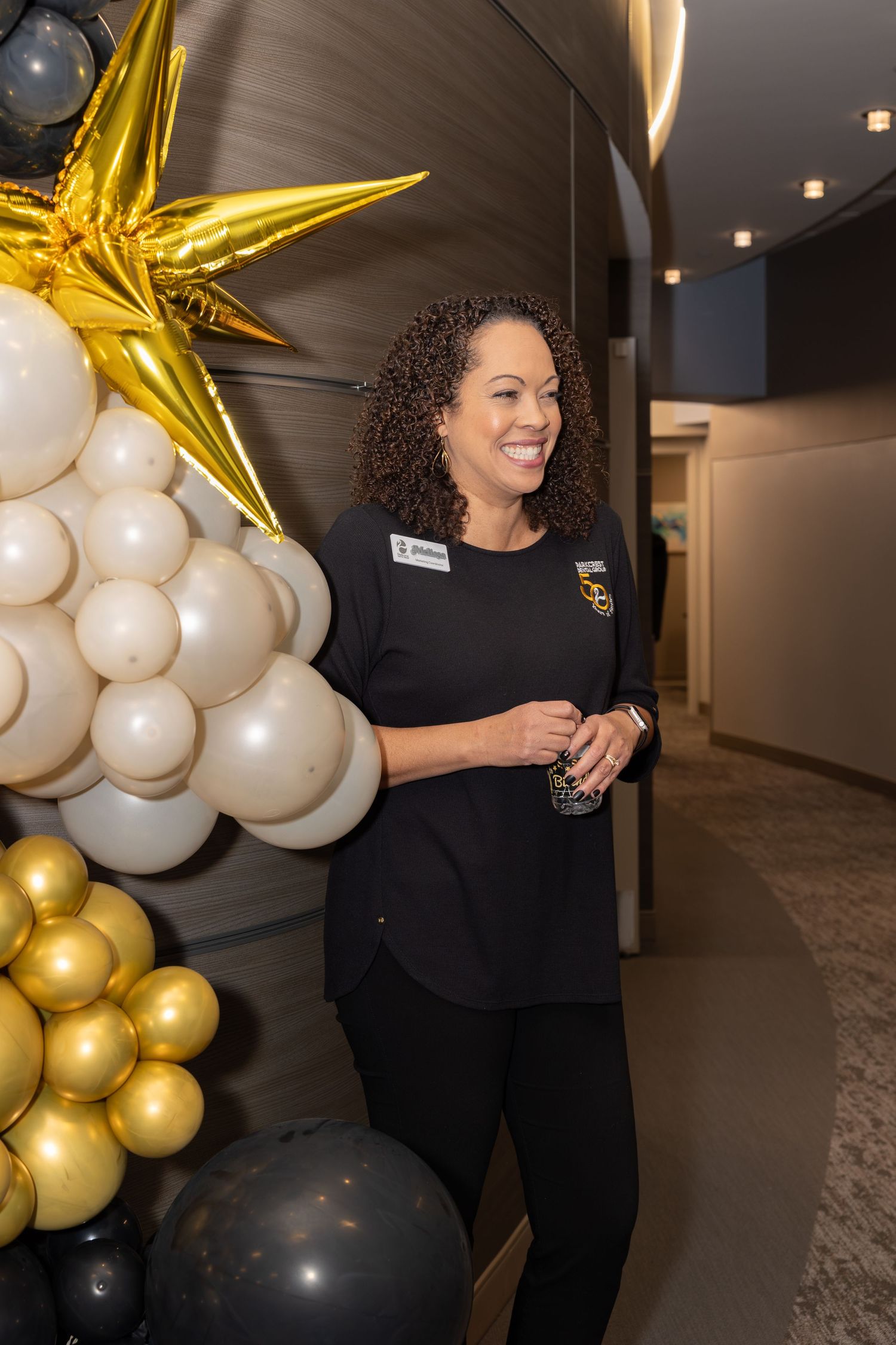 Person in black uniform smiling next to gold star and balloon decorations at an event.