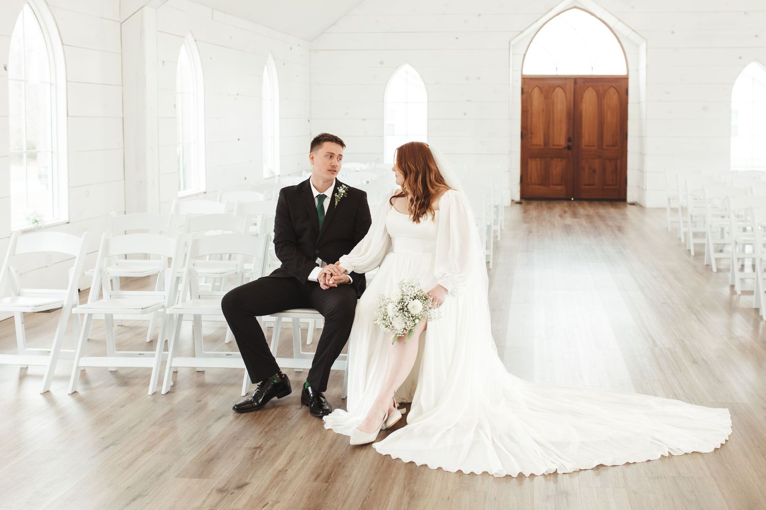 Wedding couple seated in an elegant white chapel with arched windows and wooden doors.