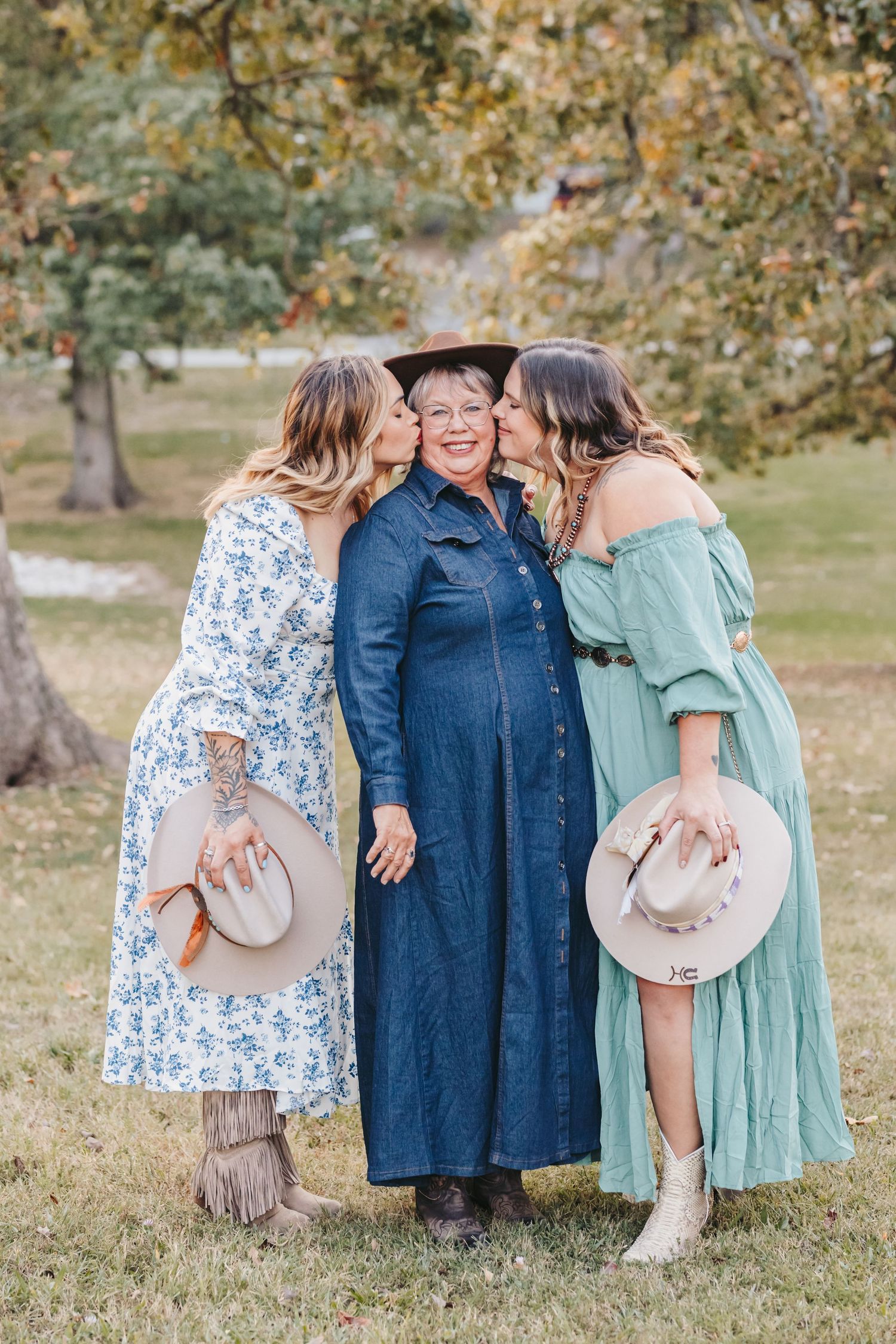 Three people share an affectionate moment outdoors wearing cowboy hats and flowing dresses.