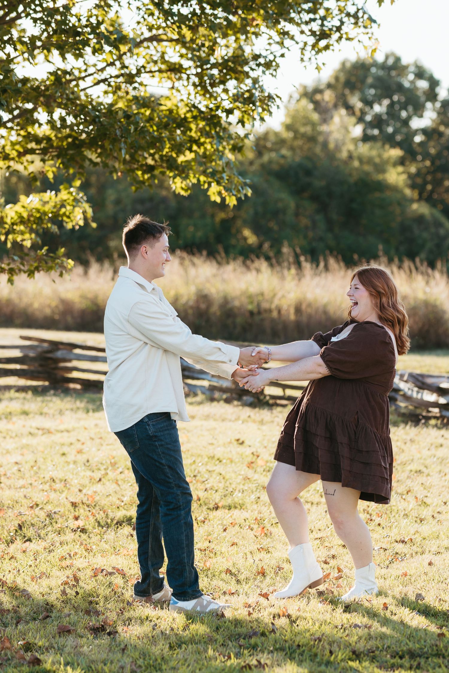 A couple dances playfully together in a sunlit autumn field with golden leaves and wooden fencing.