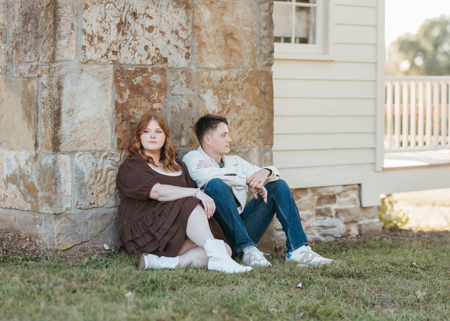 A couple sits together against a rustic stone wall of a white building.