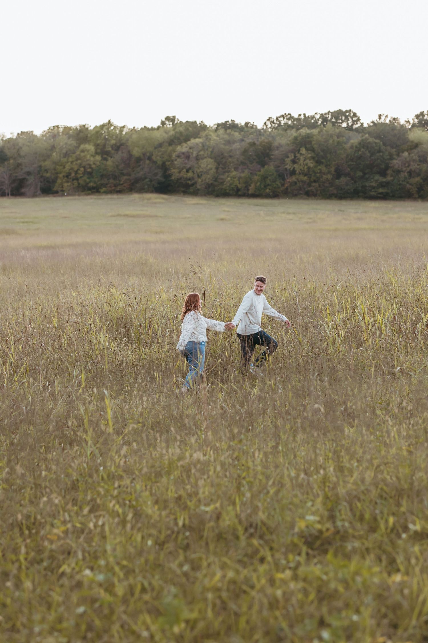 Couple strolling hand in hand through a serene meadow with treeline in background.