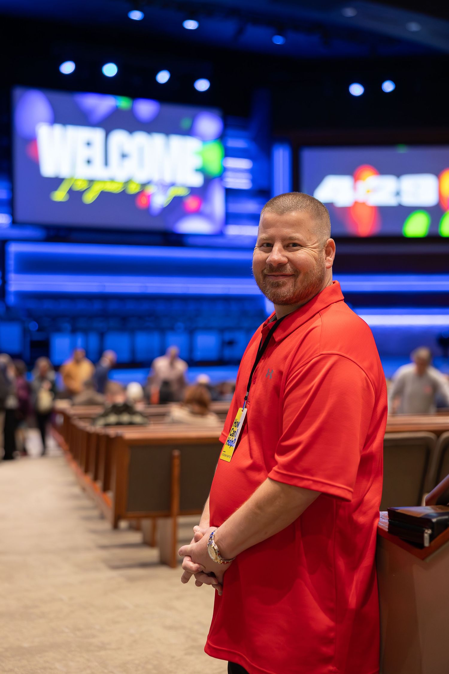 A greeter in a red polo shirt stands smiling in a church auditorium with welcome signs.