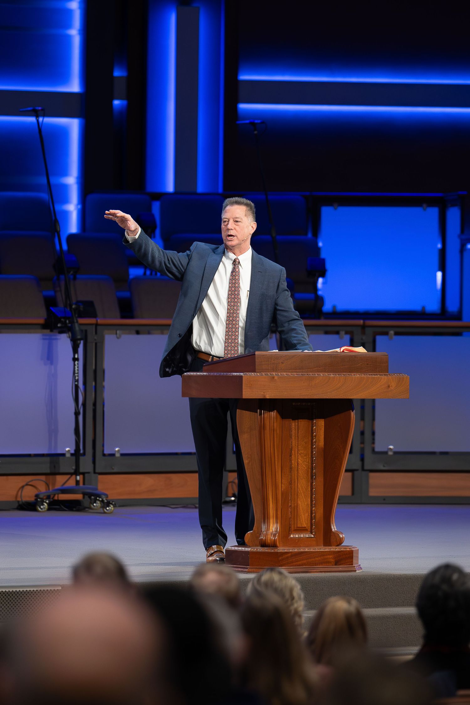 A speaker delivers a presentation from a wooden podium on stage with blue accent lighting in the background.