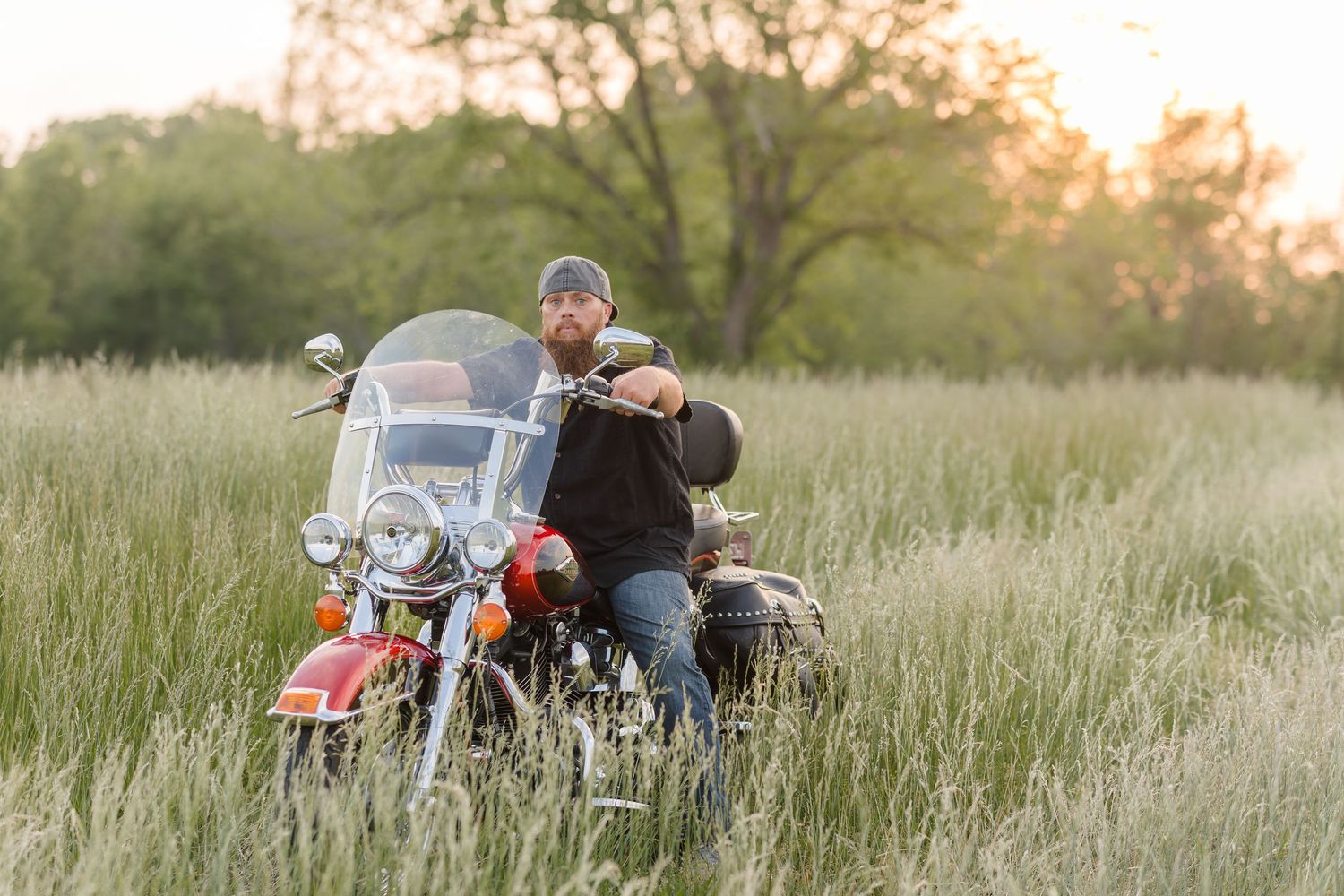 A couple sits on a parked motorcycle in a sunlit meadow of tall grass during golden hour.