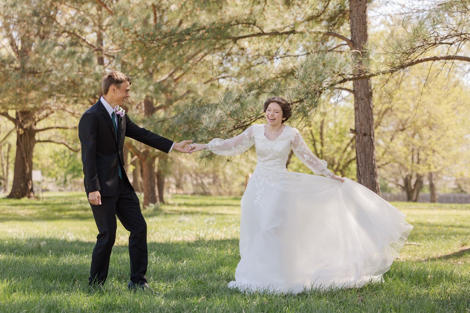 A couple in formal wedding attire shares a romantic moment in a sunlit meadow surrounded by trees and autumn foliage.