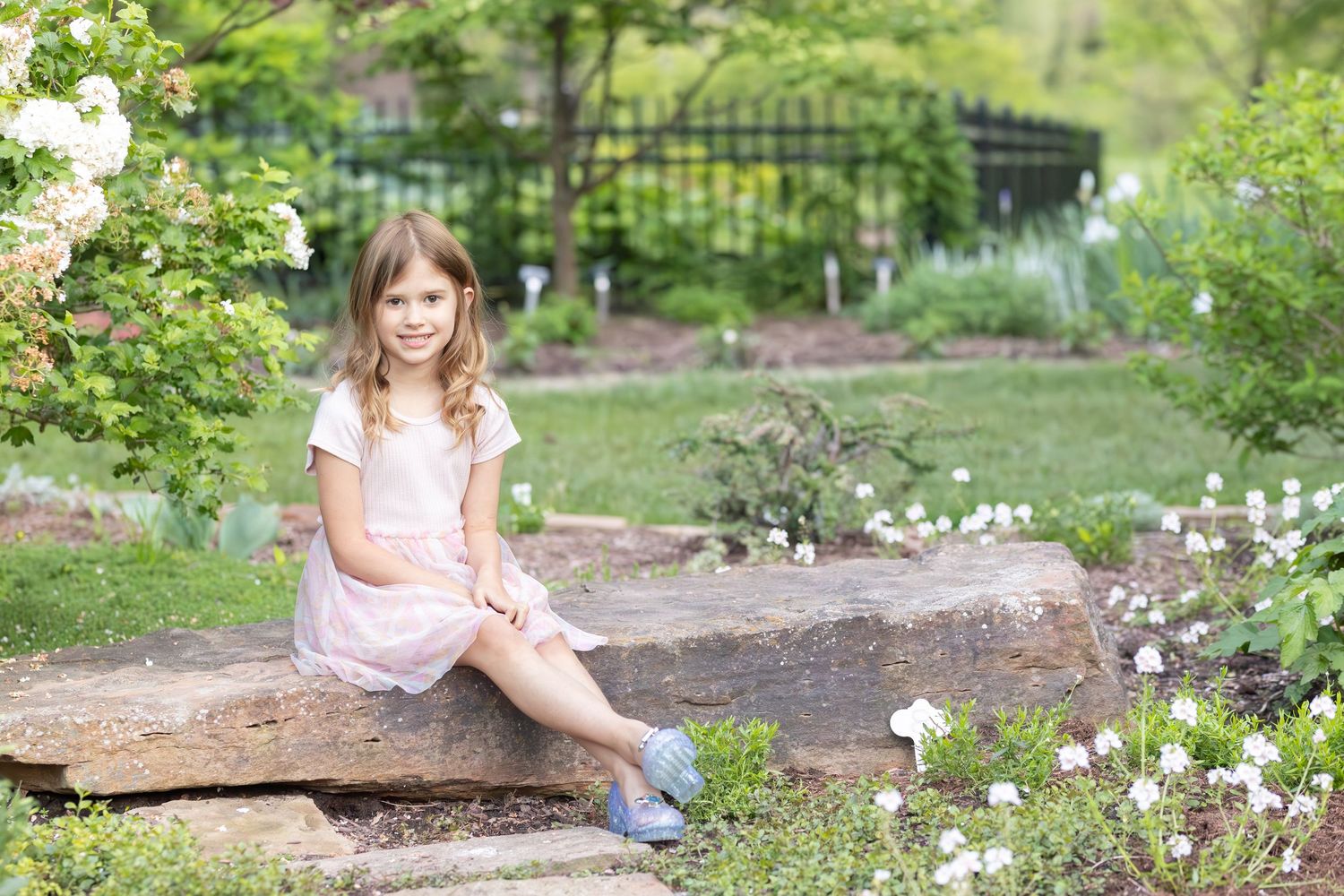 Child sitting on large rock in garden setting surrounded by spring flowers and greenery in soft natural light.