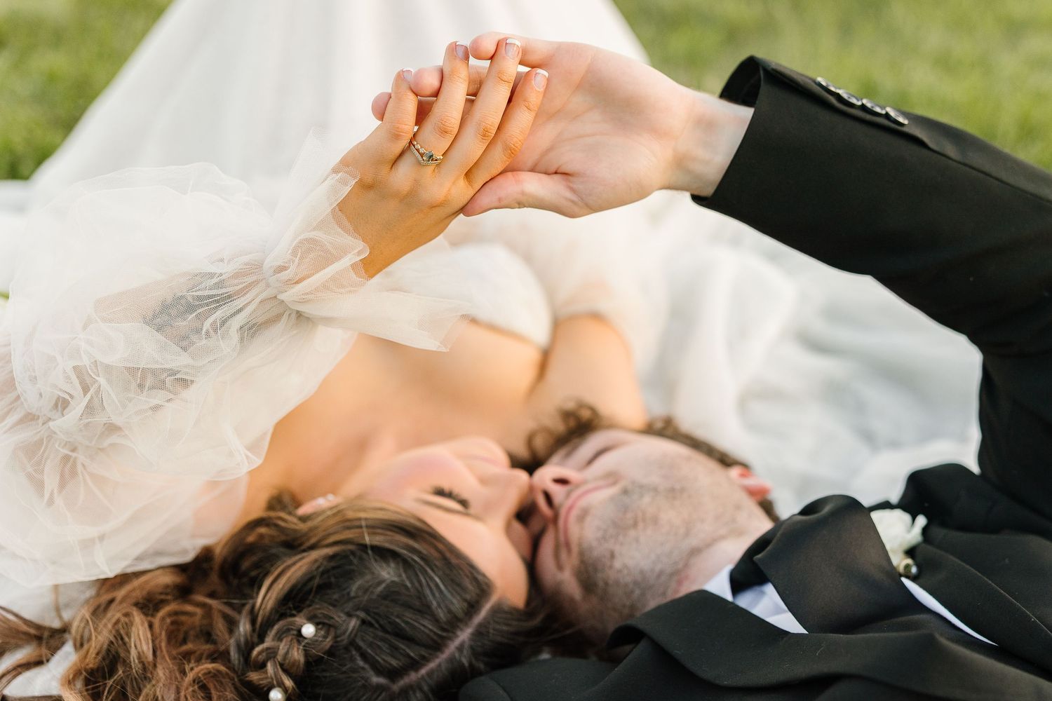 A bride and groom lay together holding hands while wearing wedding attire with a veil flowing beside them.