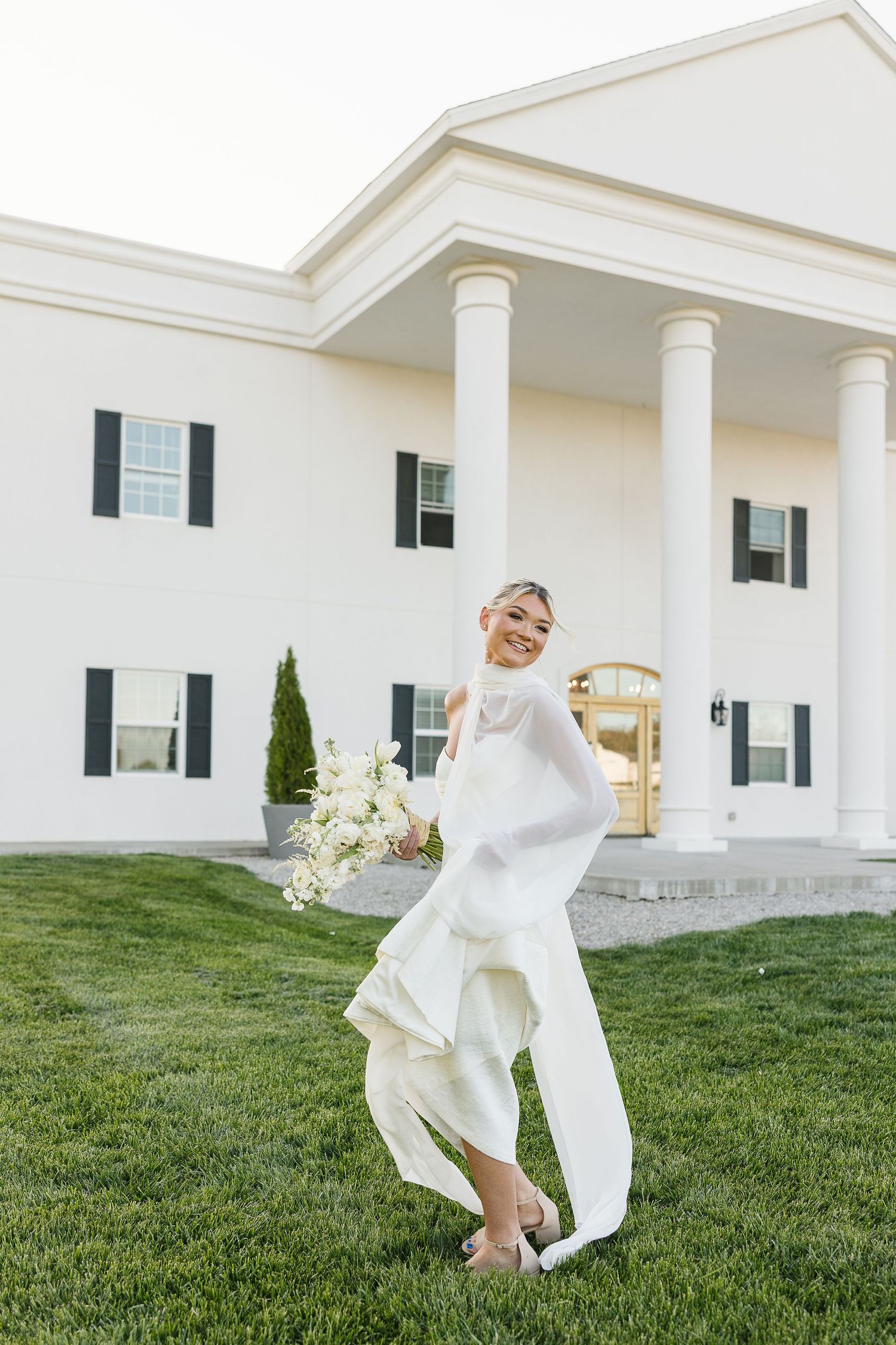 A flowing white wedding dress twirls gracefully on a green lawn in front of a stately white columned building.