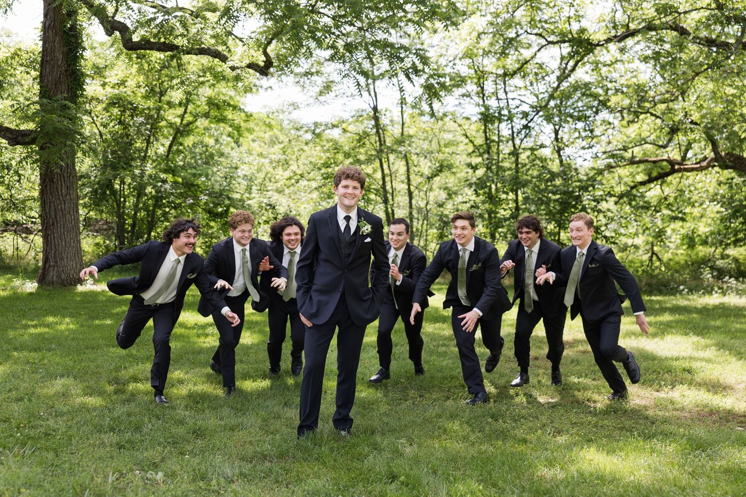 Groom and groomsmen in black suits laughing and jumping outdoors in a lush green park during a wedding celebration.