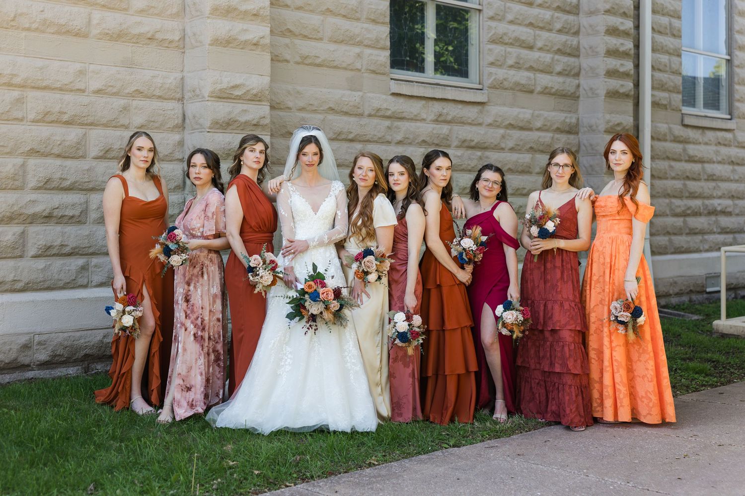 Bride in white gown poses with nine bridesmaids in mismatched rust and terracotta dresses holding bouquets outdoors.