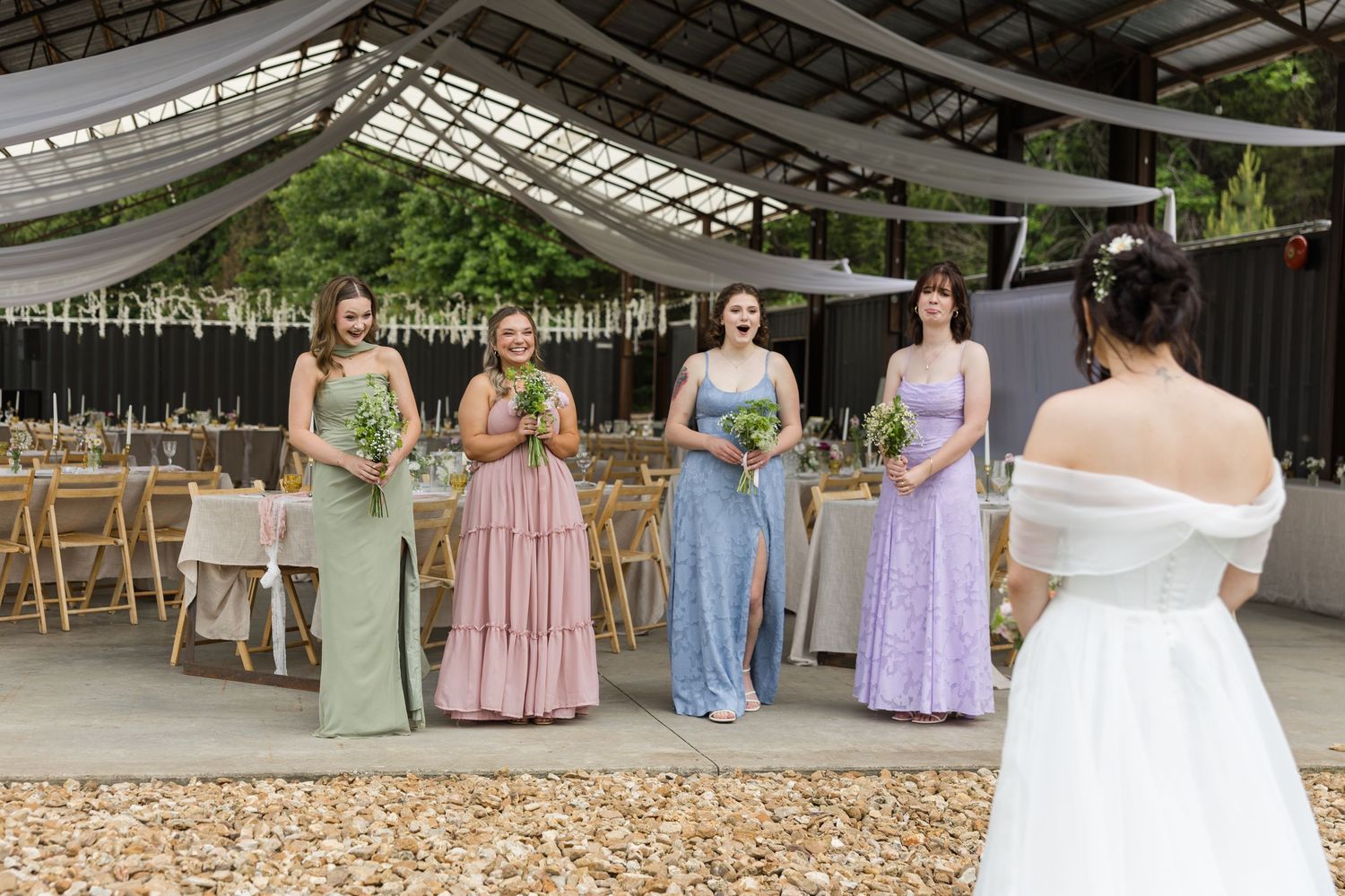 Four bridesmaids in colorful gowns holding bouquets stand before a bride in white at an elegant outdoor wedding venue.