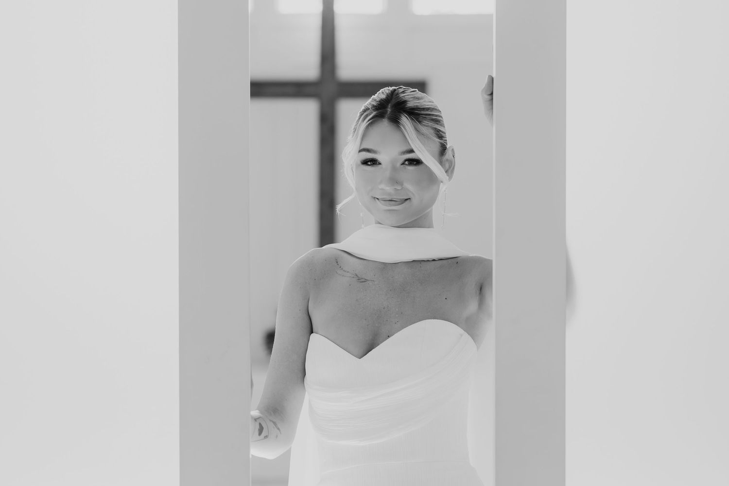 Black and white portrait of a smiling bride in a strapless wedding gown standing in a doorway with a cross in background.