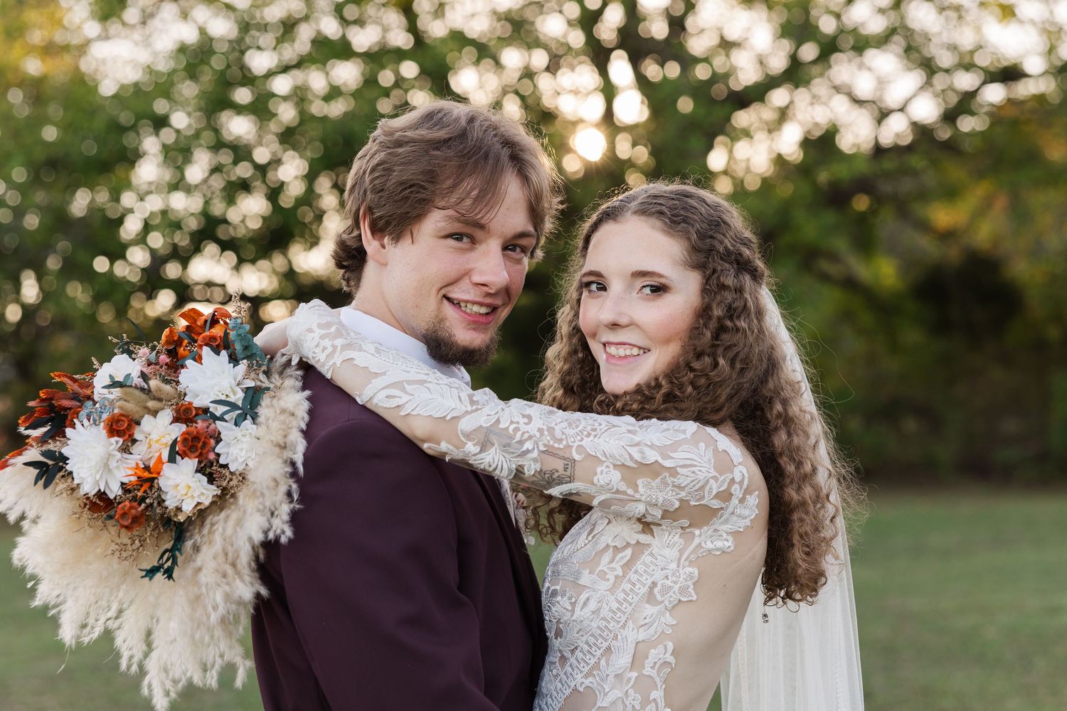 Bride and groom posing outdoors, she holds a boho bouquet with rust flowers, wearing lace gown, he in burgundy suit.