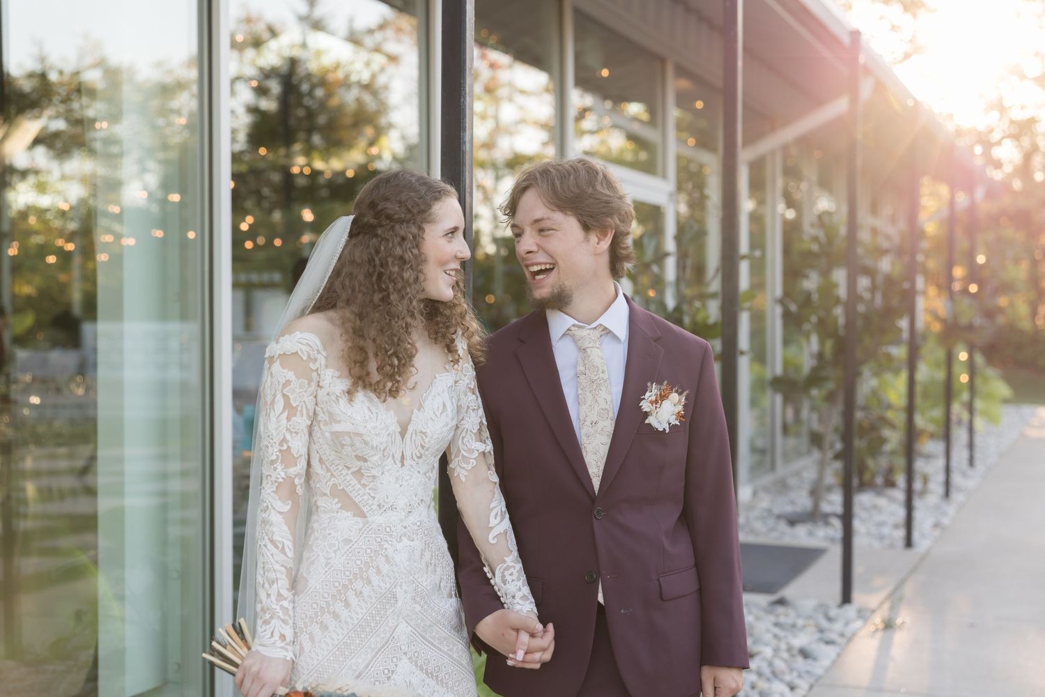 Smiling bride and groom hold hands in golden hour sunlight outside a modern glass building.