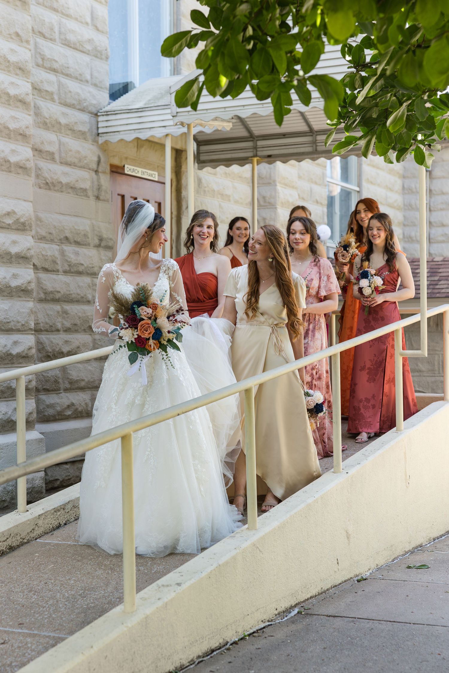 Bride in white lace gown holds bouquet with bridesmaids in red and gold dresses on church steps.