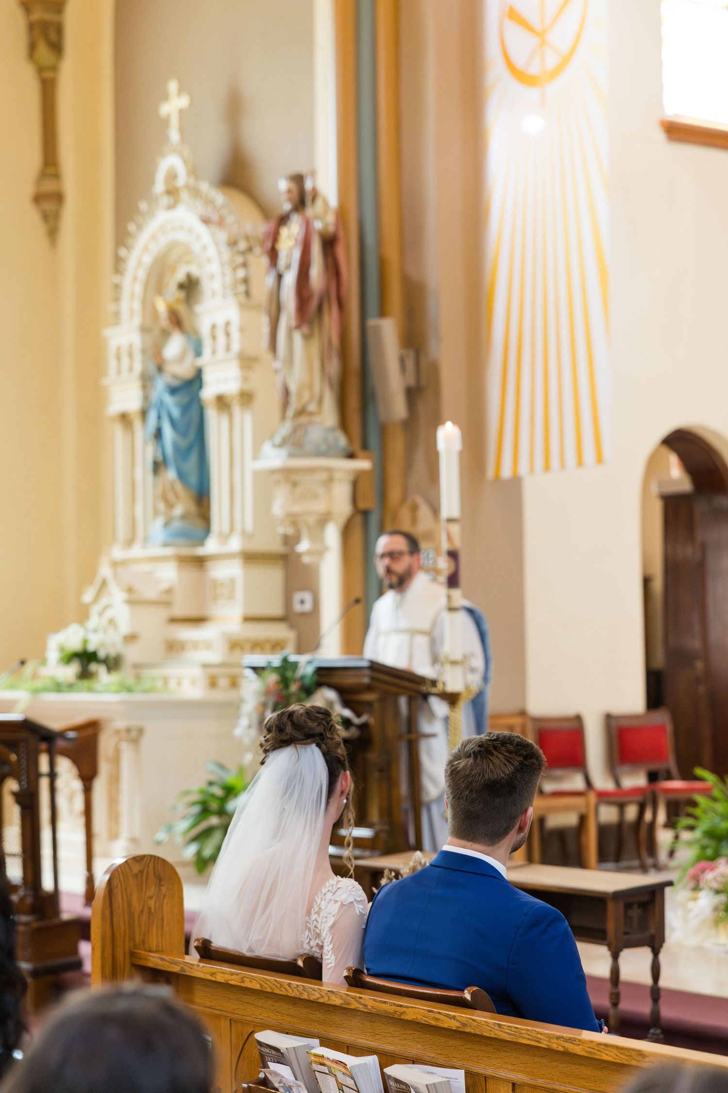 Bride and groom kneel at church altar during Catholic wedding ceremony with priest officiating.