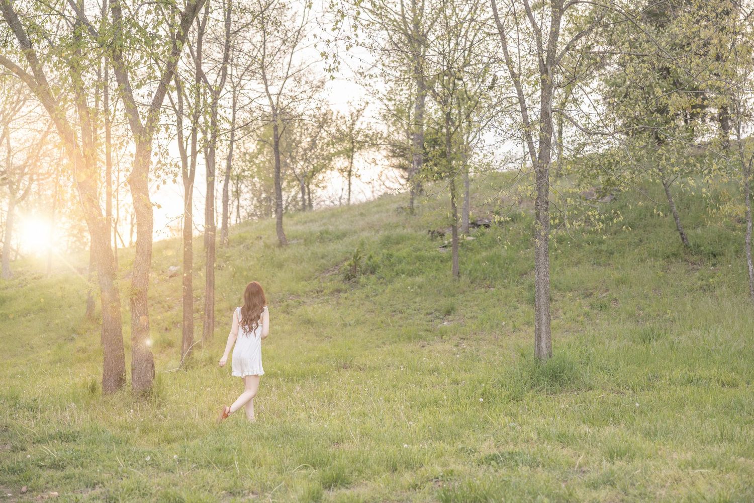 A figure in a flowy white dress walks through a sunlit hillside meadow among scattered trees during golden hour.