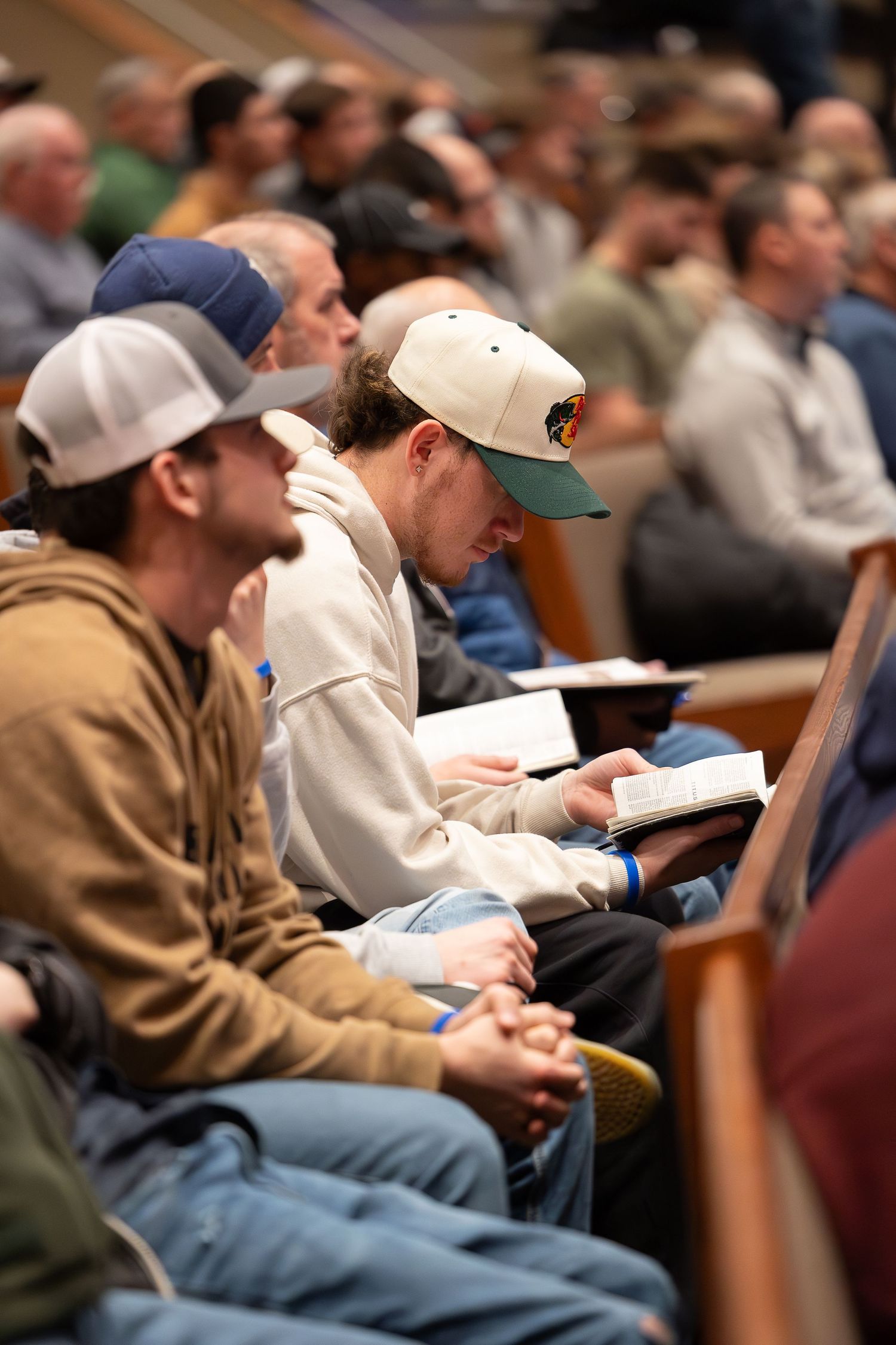Students take notes during a lecture in a large university classroom while wearing casual attire and baseball caps.