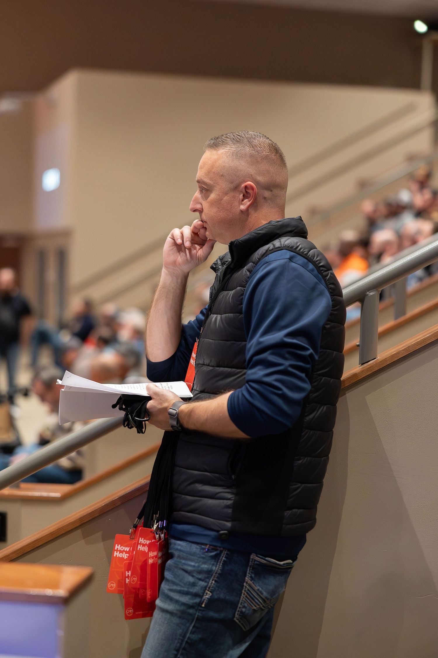 A person in a navy puffer vest and jeans stands pensively in an auditorium while holding papers and a red water bottle.