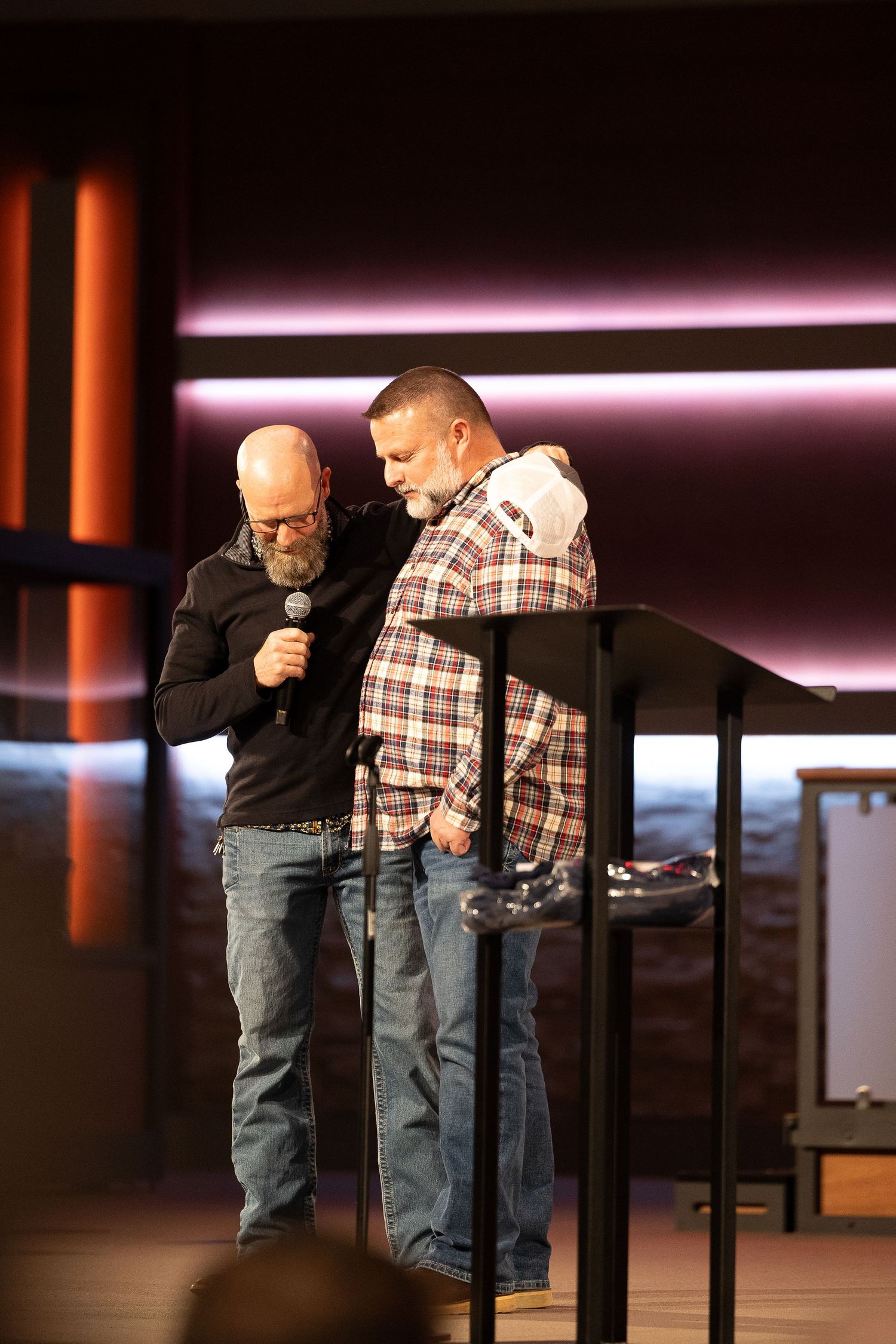 Two men stand at lecterns on a stage with purple lighting in what appears to be a church or auditorium setting.