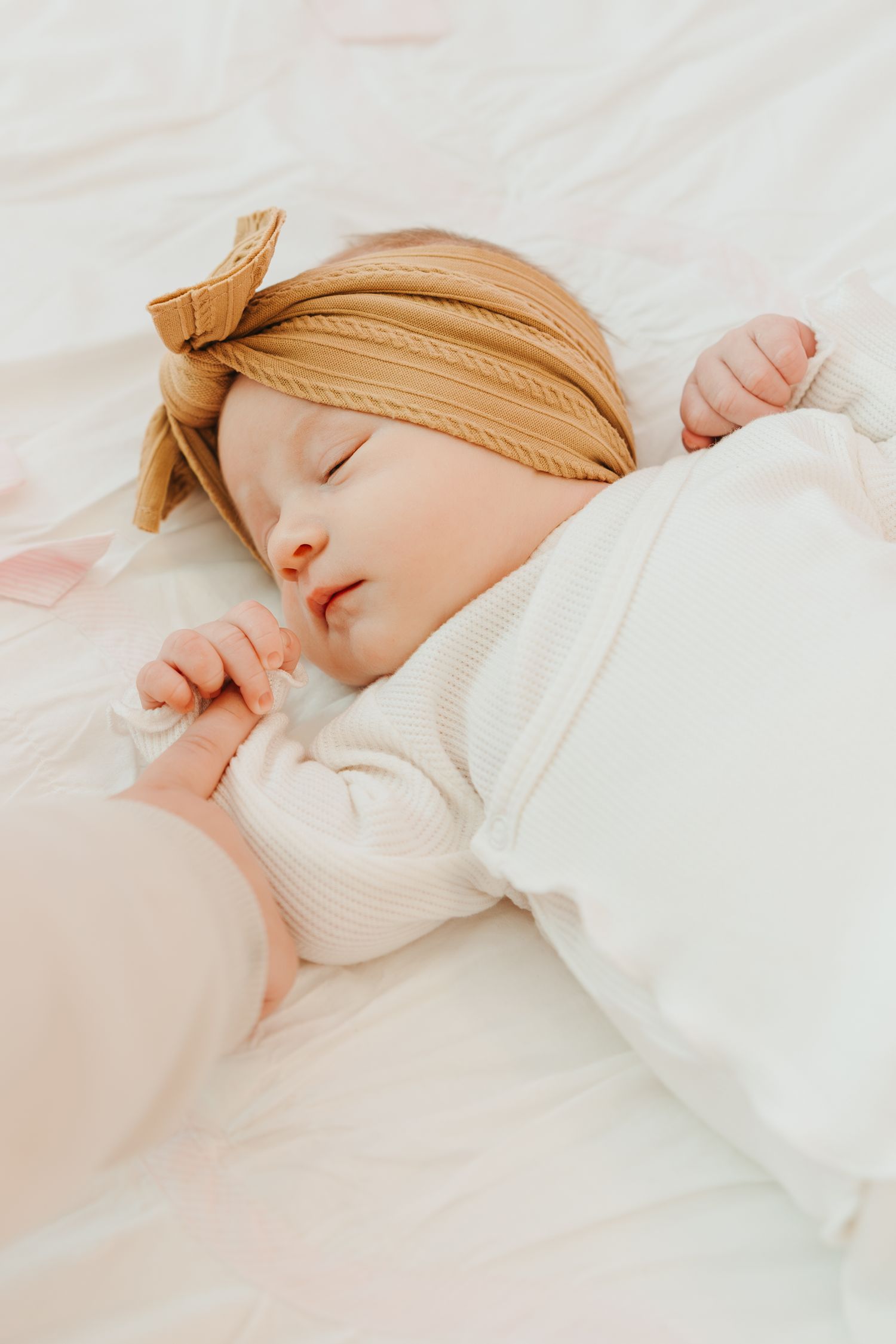 Peaceful sleeping baby wearing a mustard yellow headband and white sweater rests on soft white bedding.