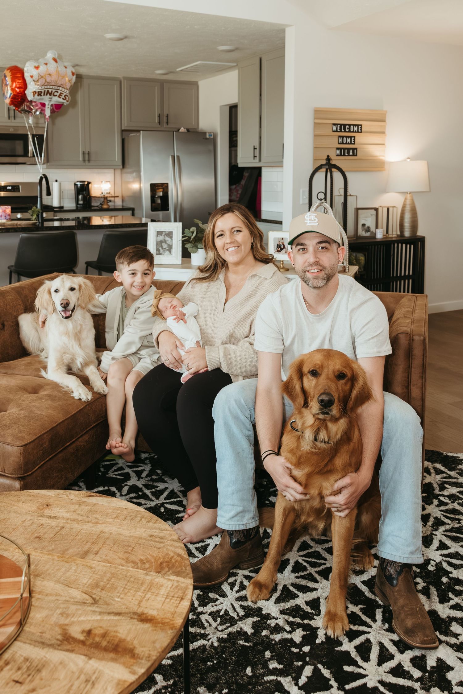 A family sits together on a brown couch with their two dogs in a modern kitchen living room setting.