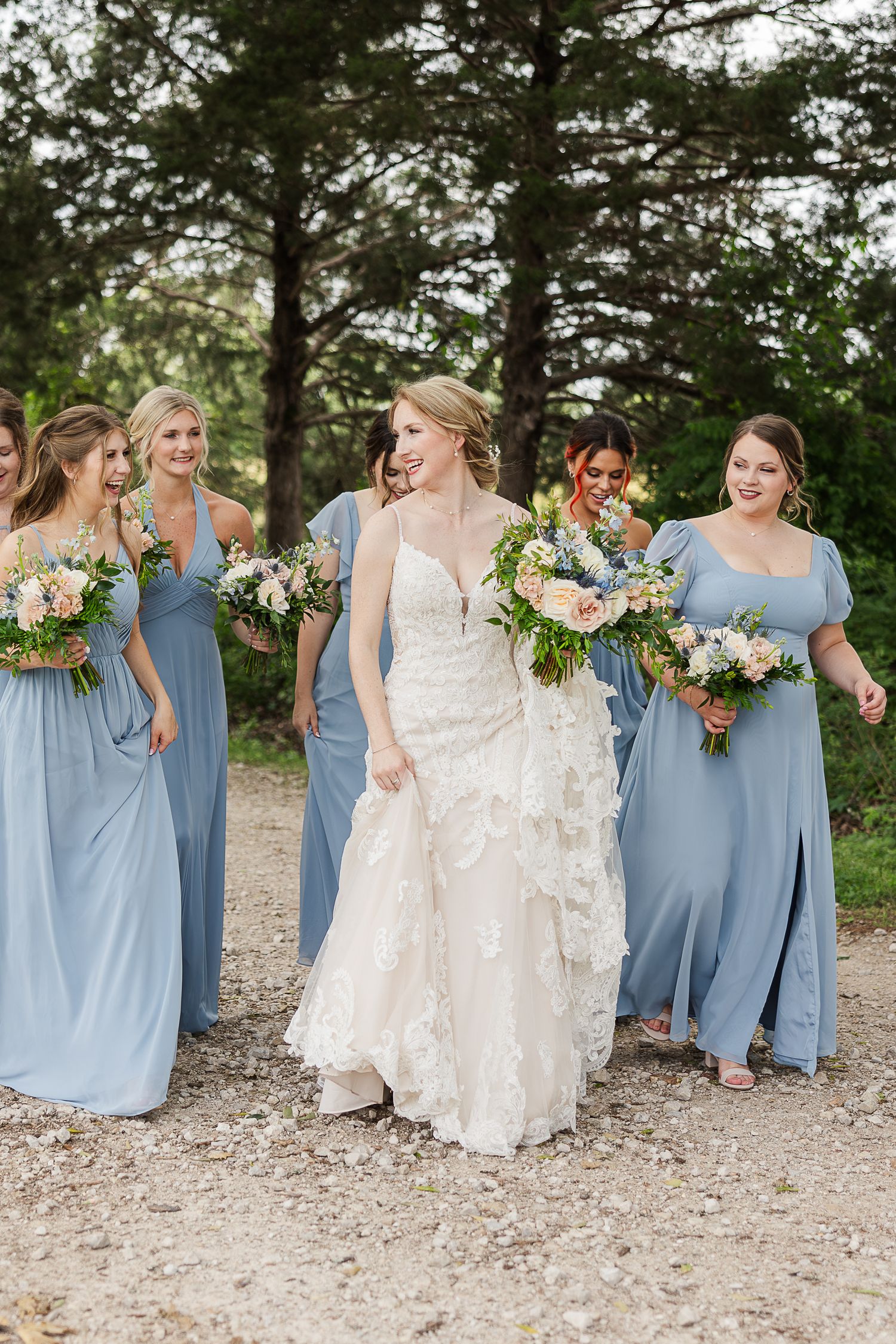 A group in pale blue bridesmaid dresses and a white wedding gown walk together outdoors holding floral bouquets.