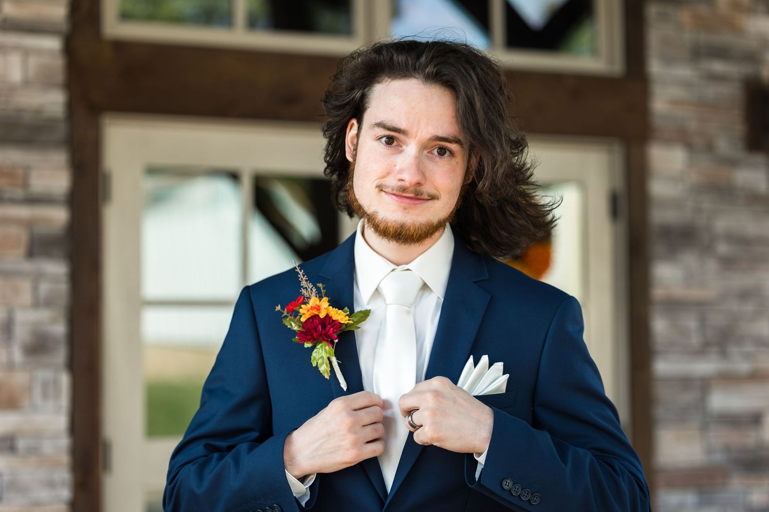 Groom in a navy blue suit adjusts his tie while standing against a brick wall.
