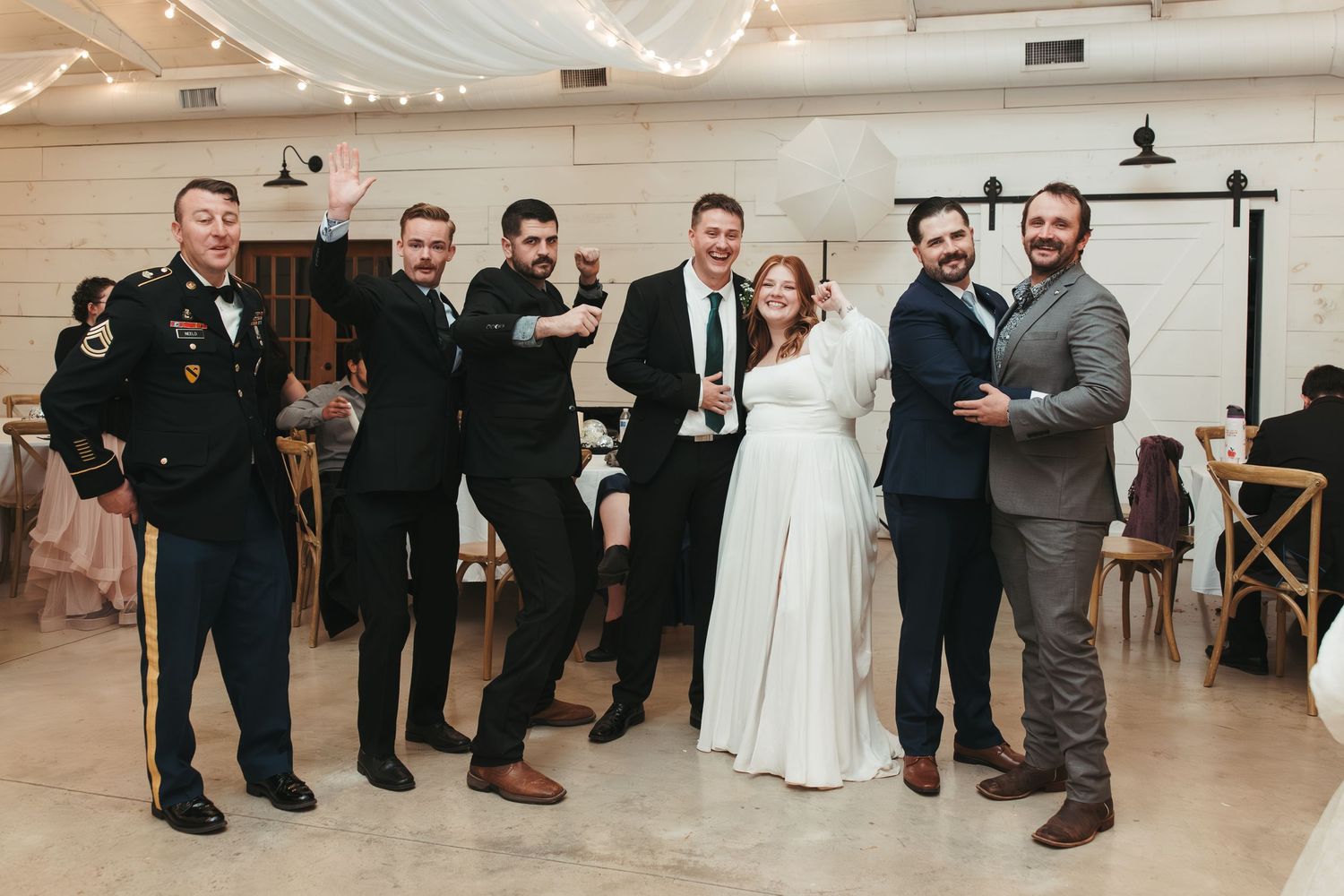 Wedding party poses in formal attire at an indoor reception venue with white walls and string lights.