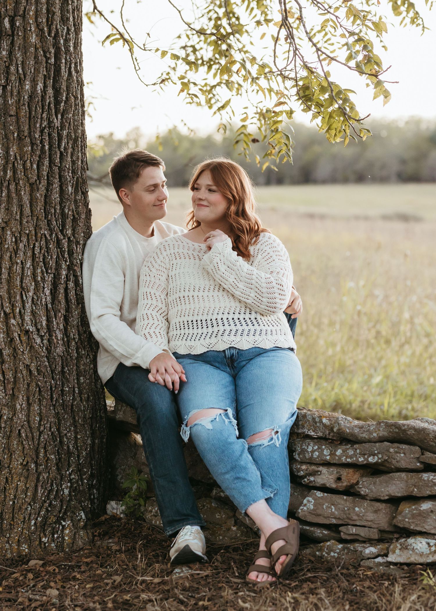 Couple in casual attire sitting together against a tree trunk in an autumn field at sunset.