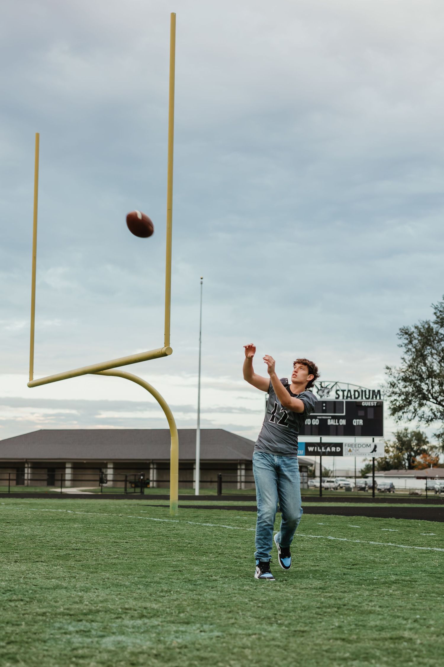 Player practices field goal kicks on empty football field with stadium scoreboard visible.