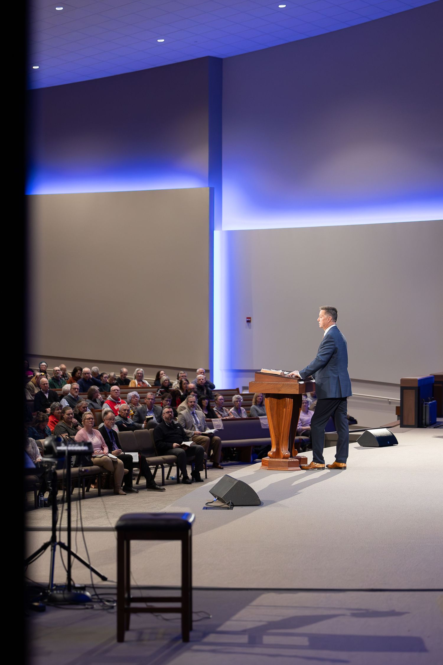 A speaker addresses a crowded auditorium from an elevated platform with modern architectural lighting.