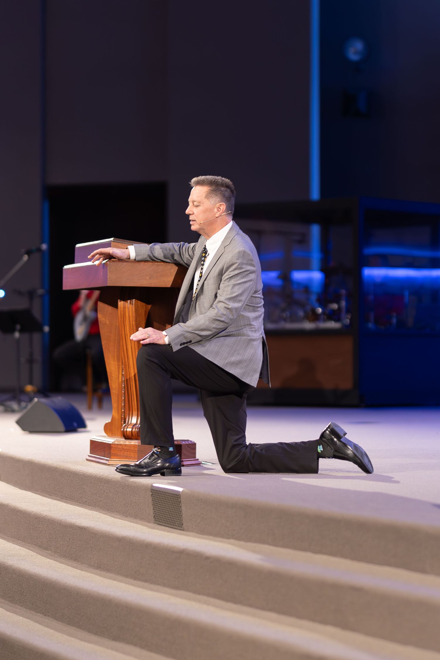 Speaker kneels at wooden podium on church stage with blue accent lighting.