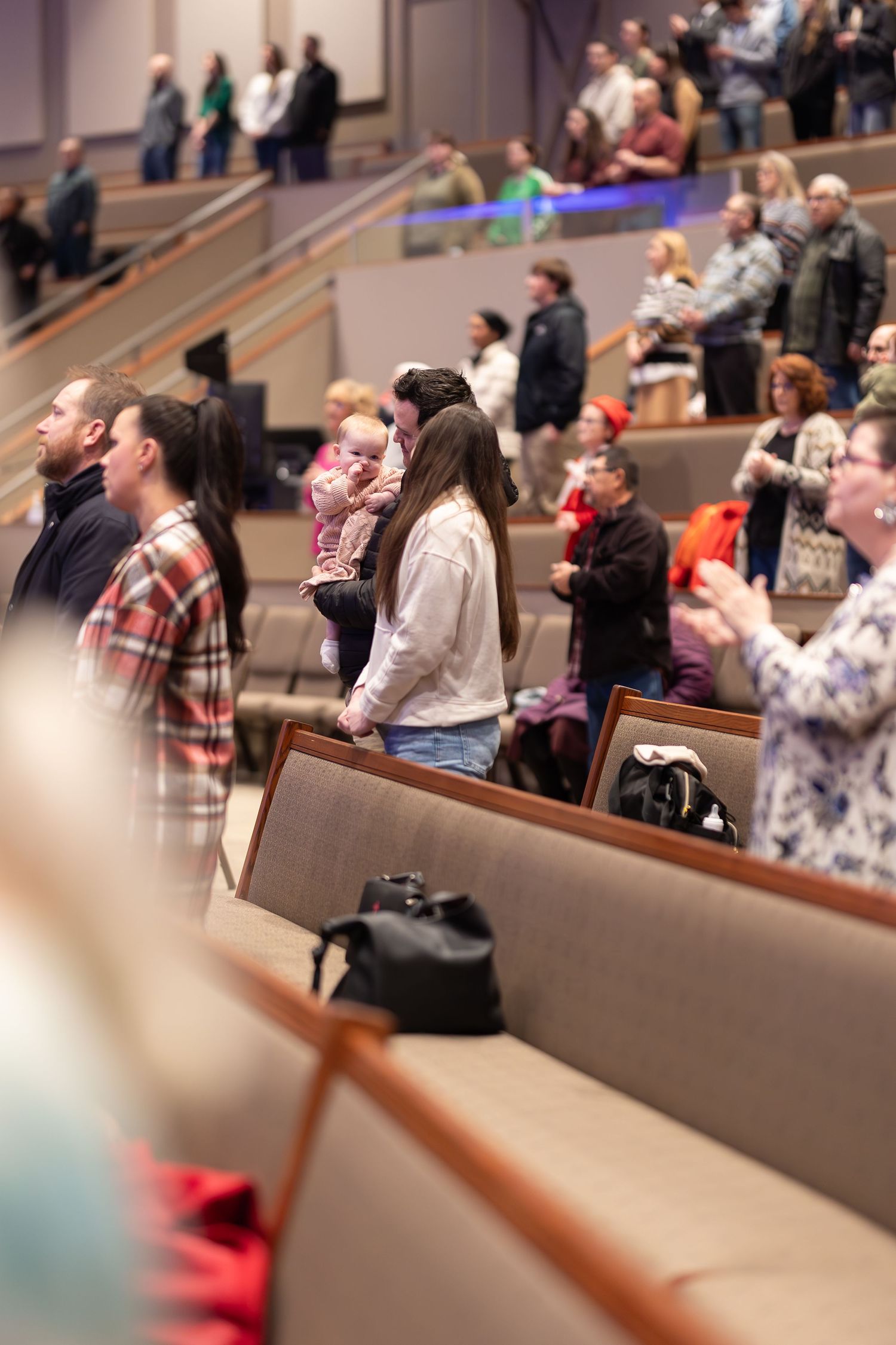 Congregation stands during worship service in modern church sanctuary with ascending rows of pews.