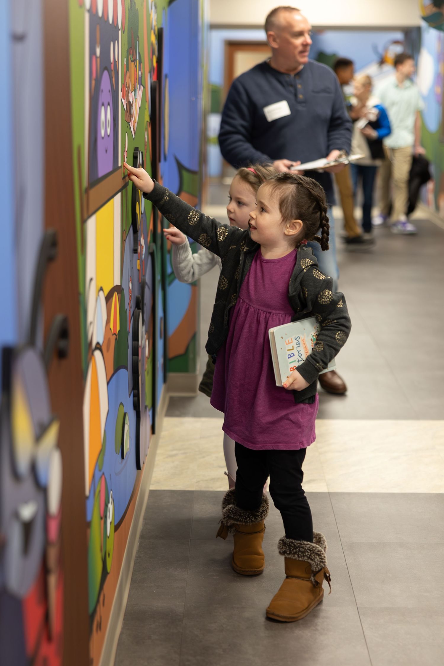 Young child in purple dress touching colorful wall display while carrying a book in school hallway.