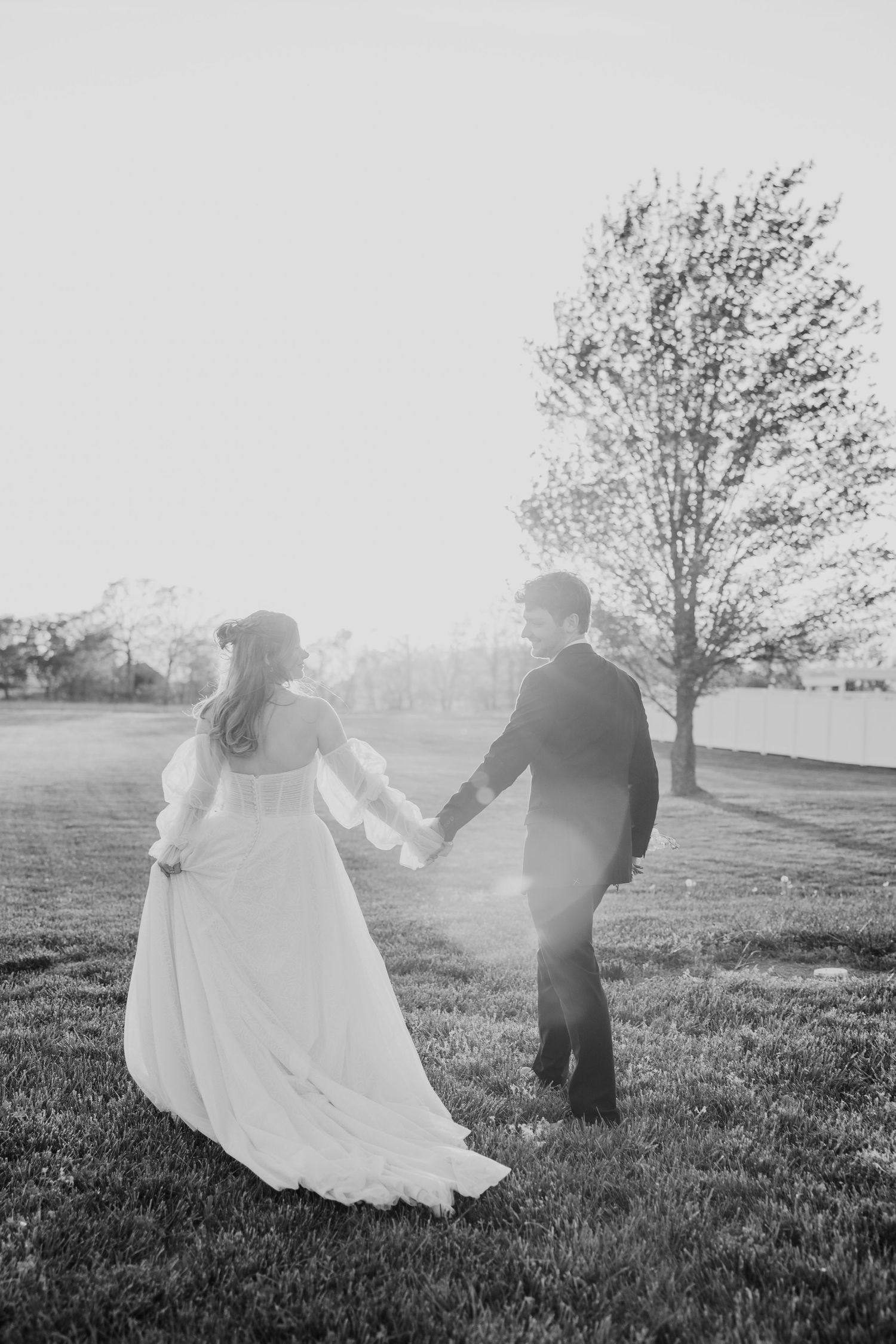 Black and white photo of bride and groom holding hands walking across a grassy field on their wedding day.