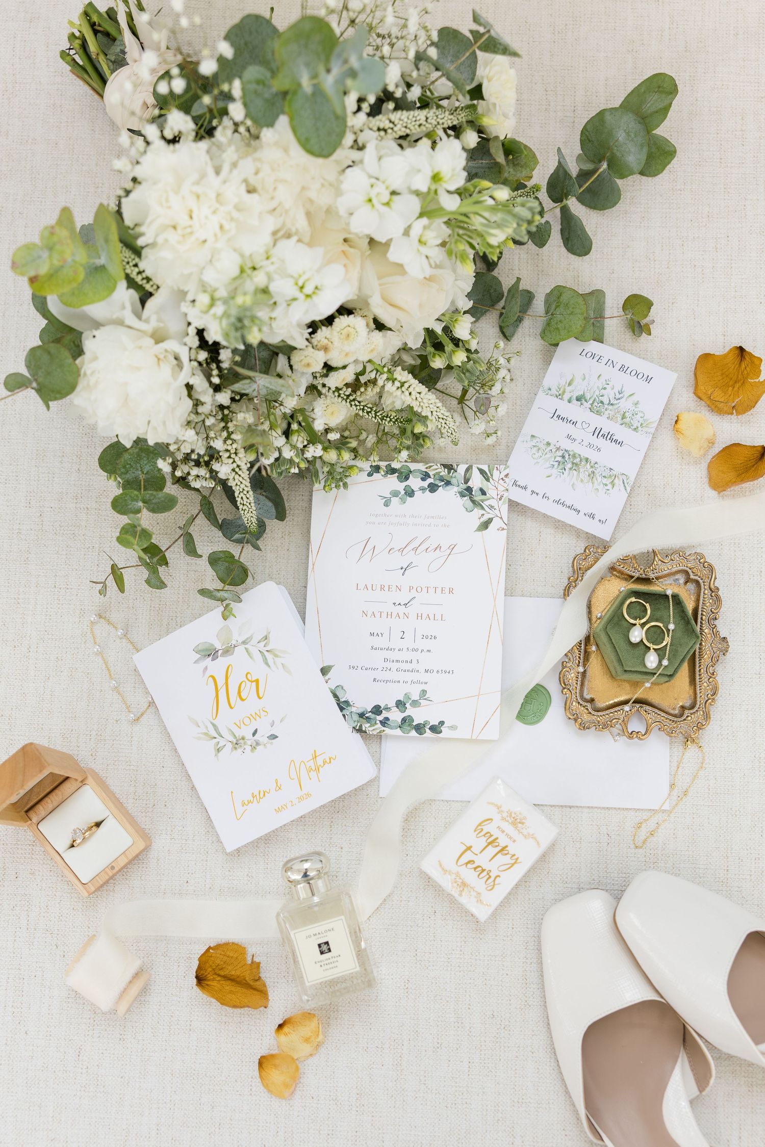 Elegant wedding flat lay with white floral bouquet, gold ring box, invitations, Jo Malone perfume, and bridal heels.