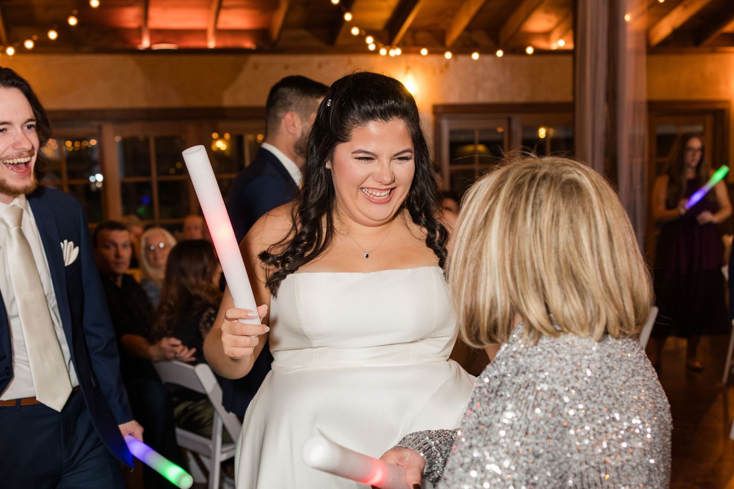 Wedding reception guests dance and celebrate under warm lighting and string lights in a rustic indoor venue.