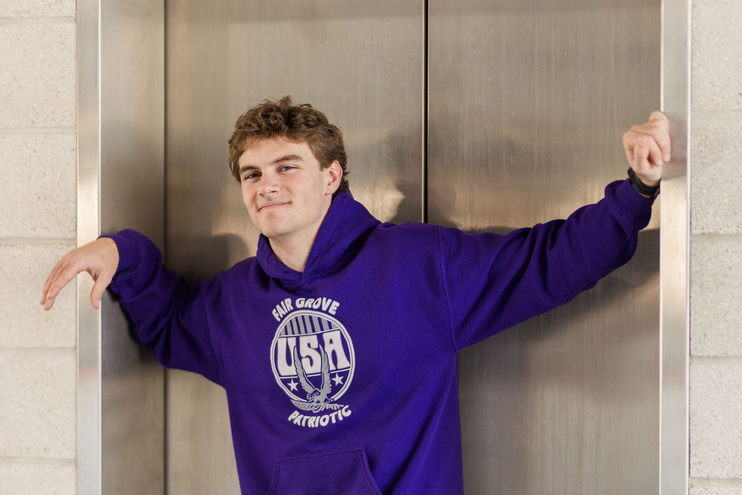 A person in a purple USA graphic sweatshirt poses against a metallic elevator door.